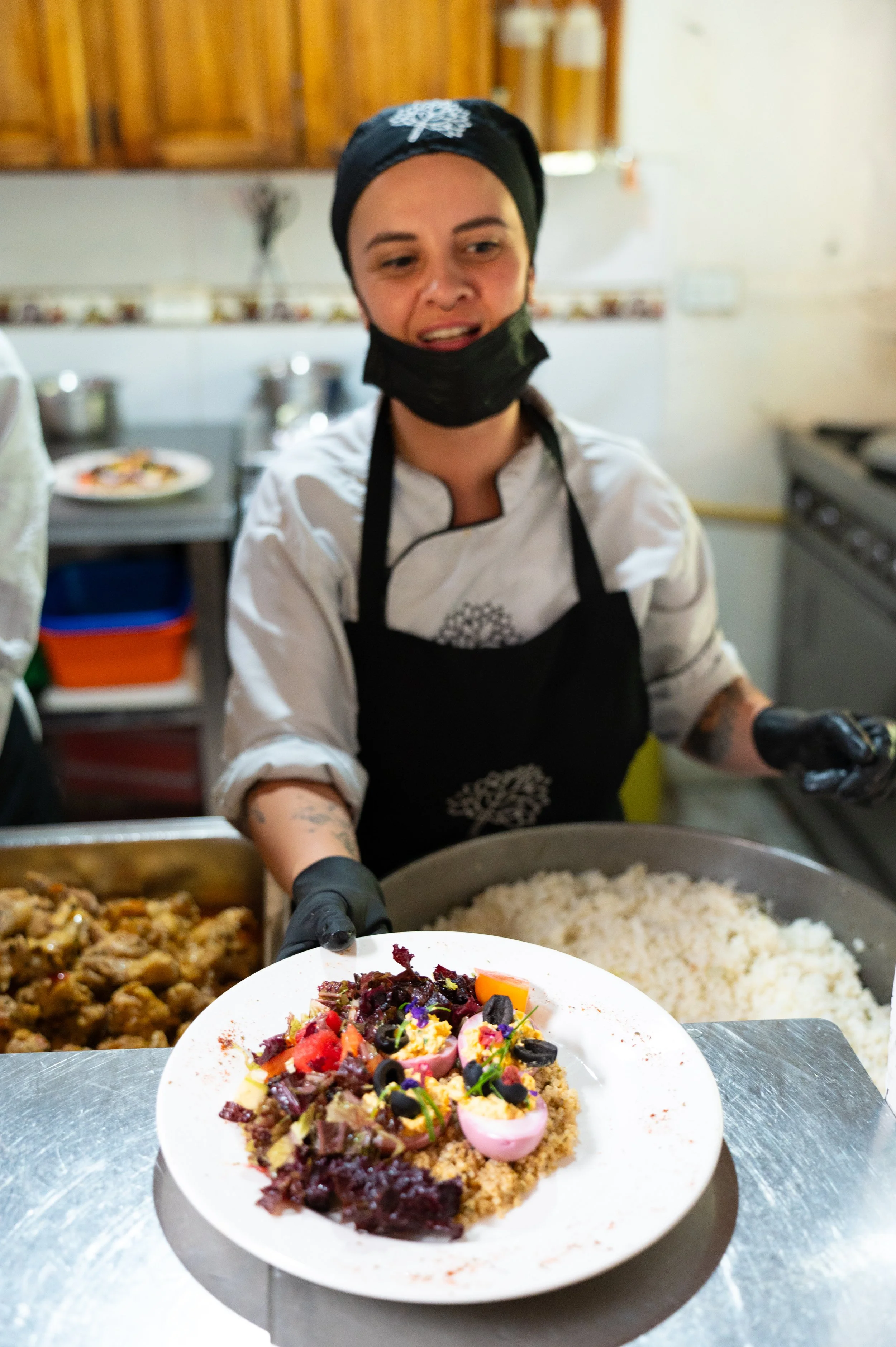 A woman wearing a black chef's hat and a black apron, working in a kitchen, preparing food with a large bowl of cooked rice and various dishes in the background, and a plate with colorful food in the foreground.