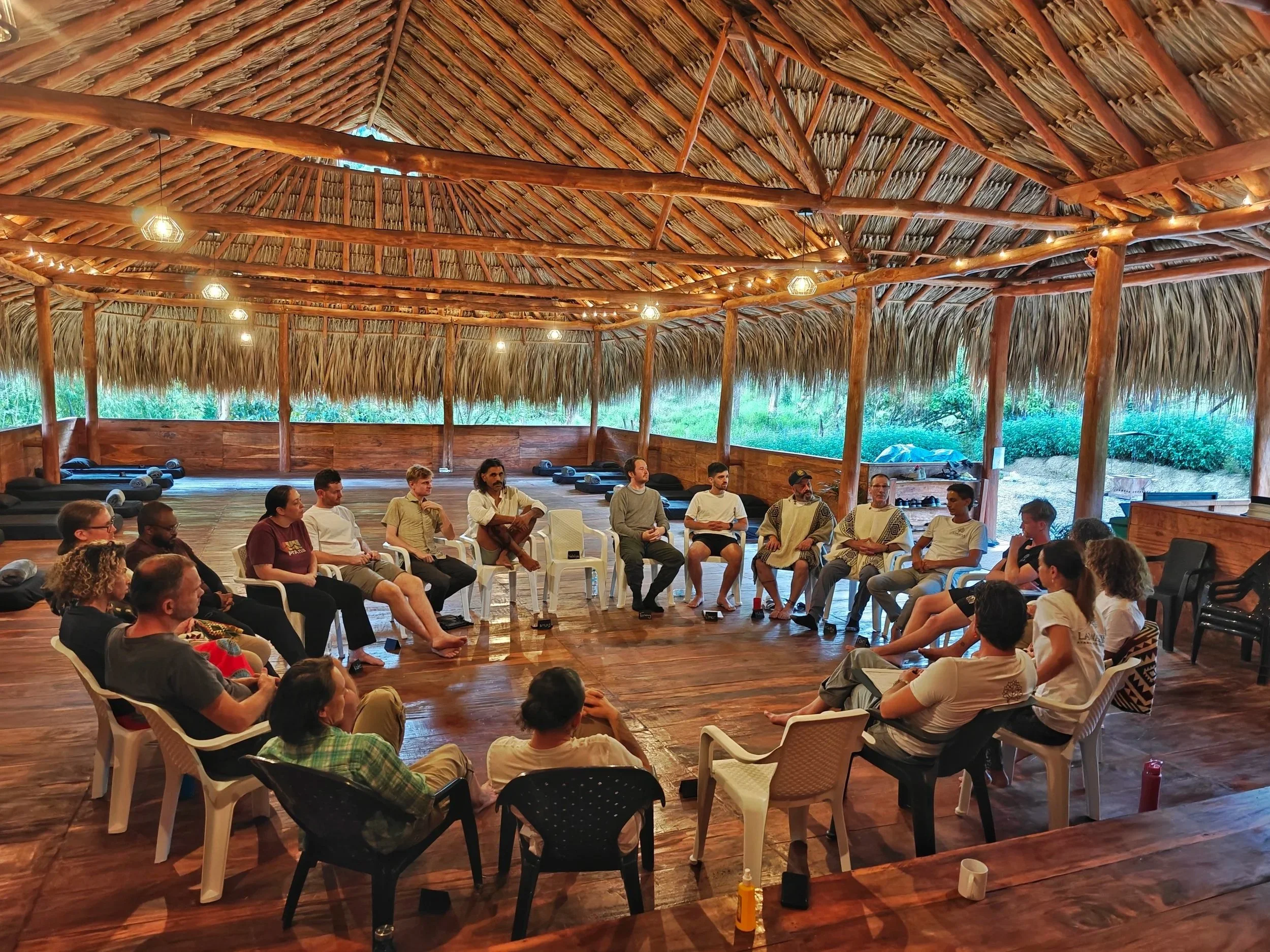 A group of people seated in a semi-circle inside a large open-sided structure with a thatched roof, participating in or attending a discussion or presentation during daylight.