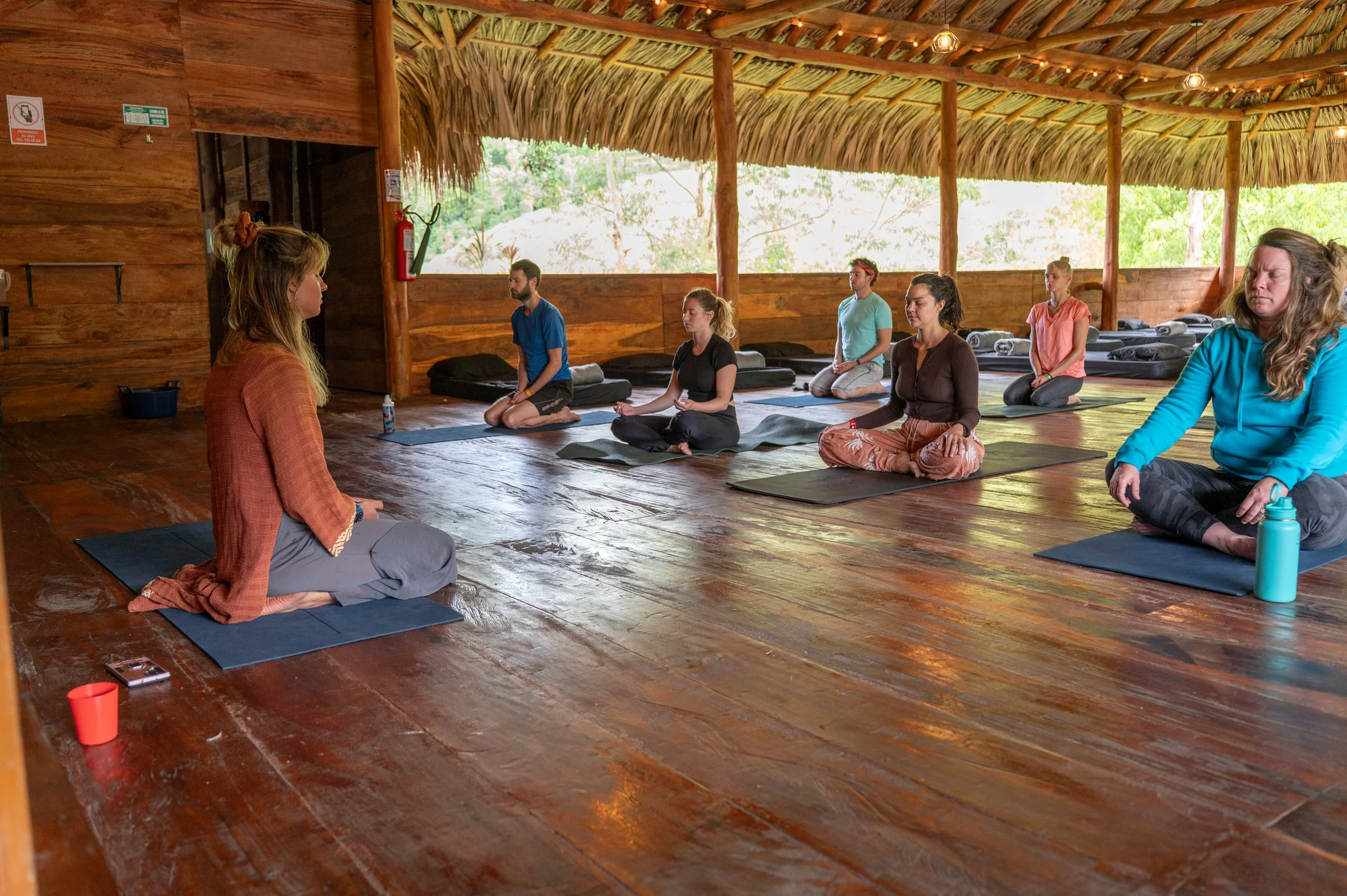 A group of people practicing yoga or meditation in a rustic, open-air wooden pavilion with a thatched roof surrounded by greenery.