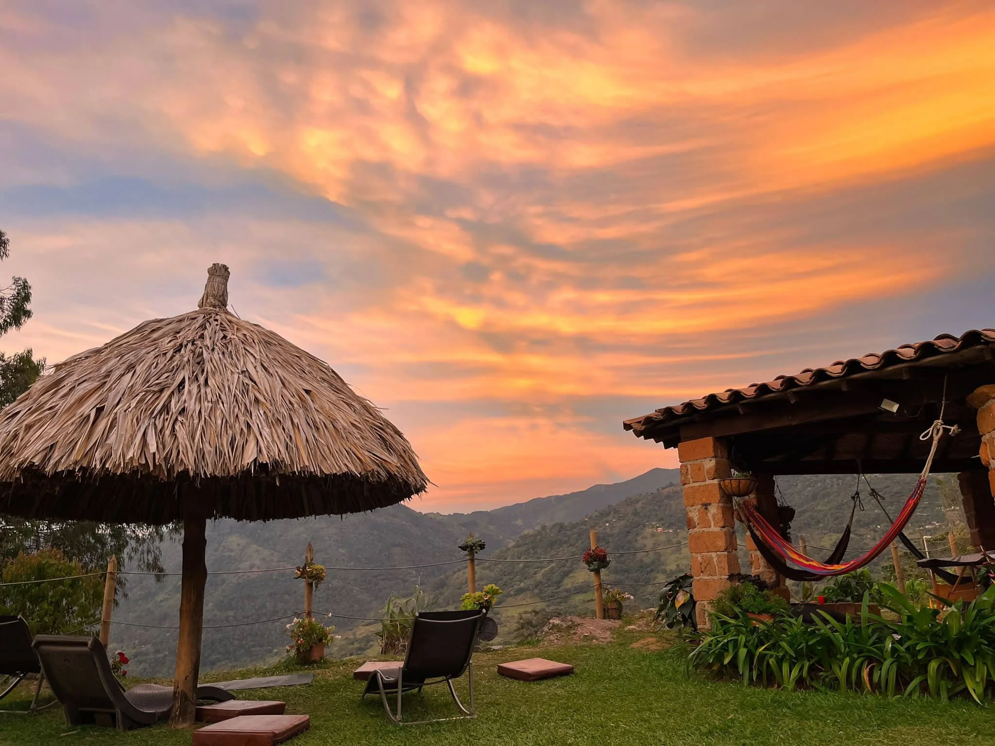 A scenic outdoor patio with a thatched umbrella, lounge chairs, potted plants, and a hammock, overlooking a mountainous landscape during sunset with colorful clouds in the sky.