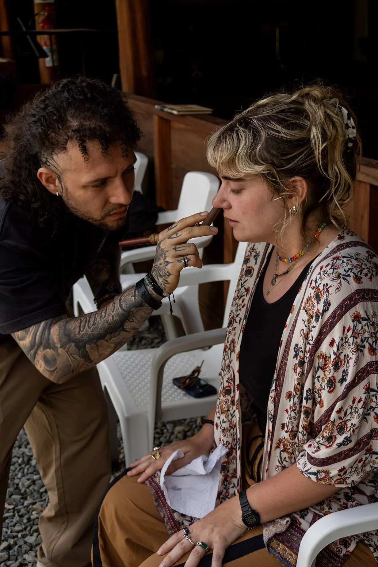 A tattooed man with curly hair and earrings applies makeup or face paint to a woman with short curly hair, wearing colorful jewelry and a patterned jacket, seated outdoors.