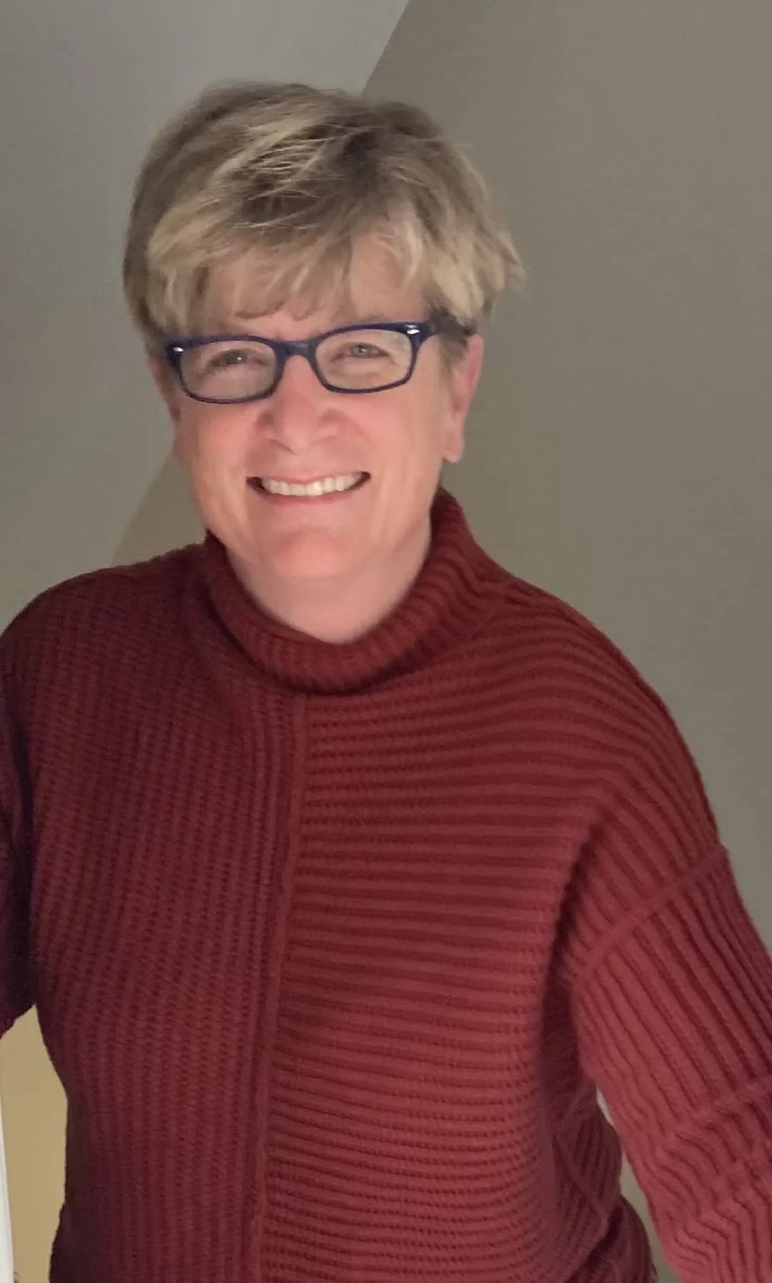 A person wearing glasses and a red, ribbed turtleneck sweater, smiling with short, light brown and blonde hair, standing against a neutral background.