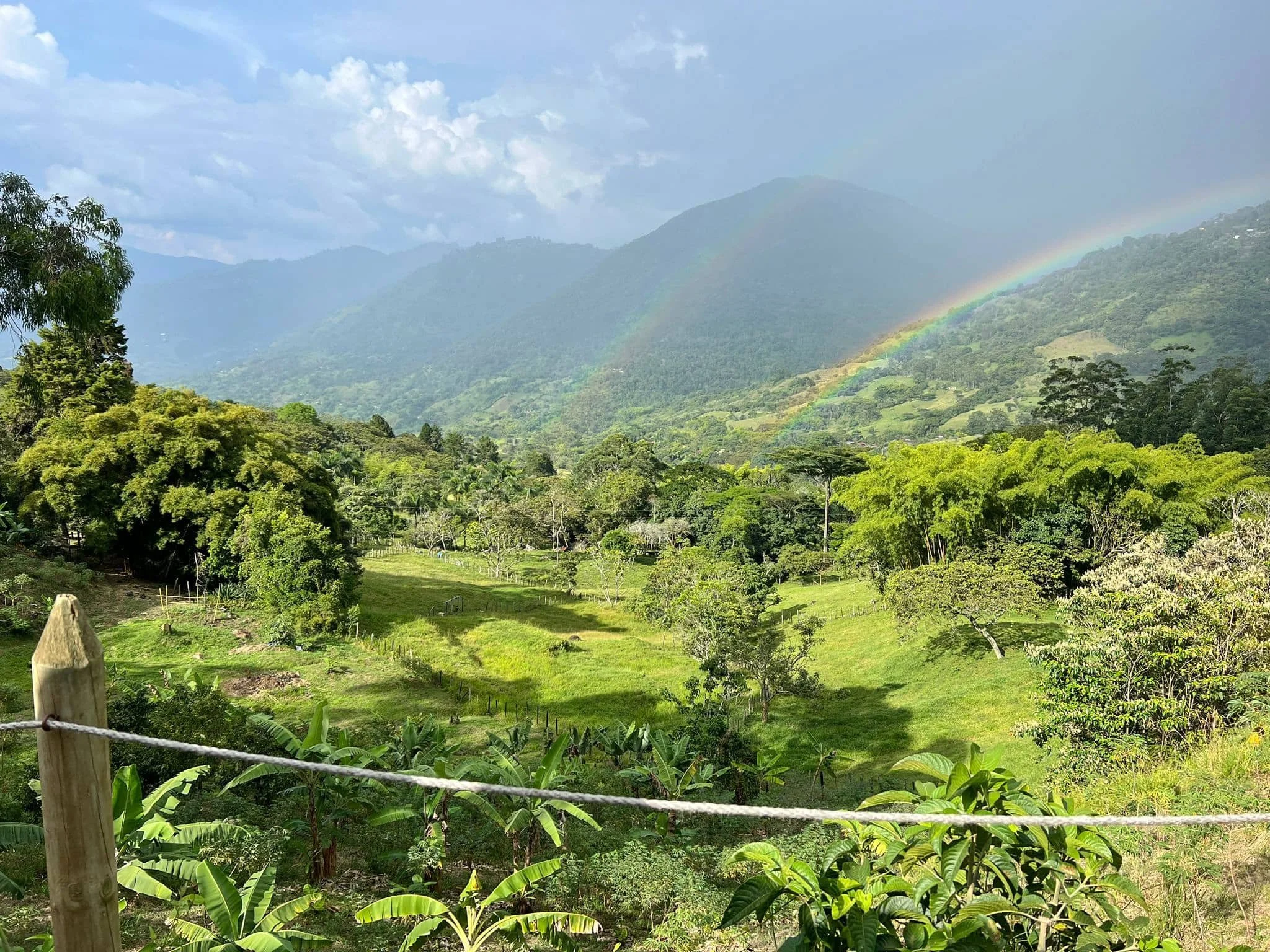 Lush green landscape with trees, grassy fields, and mountains in the background. A faint rainbow arcs across the sky, with partly cloudy weather.