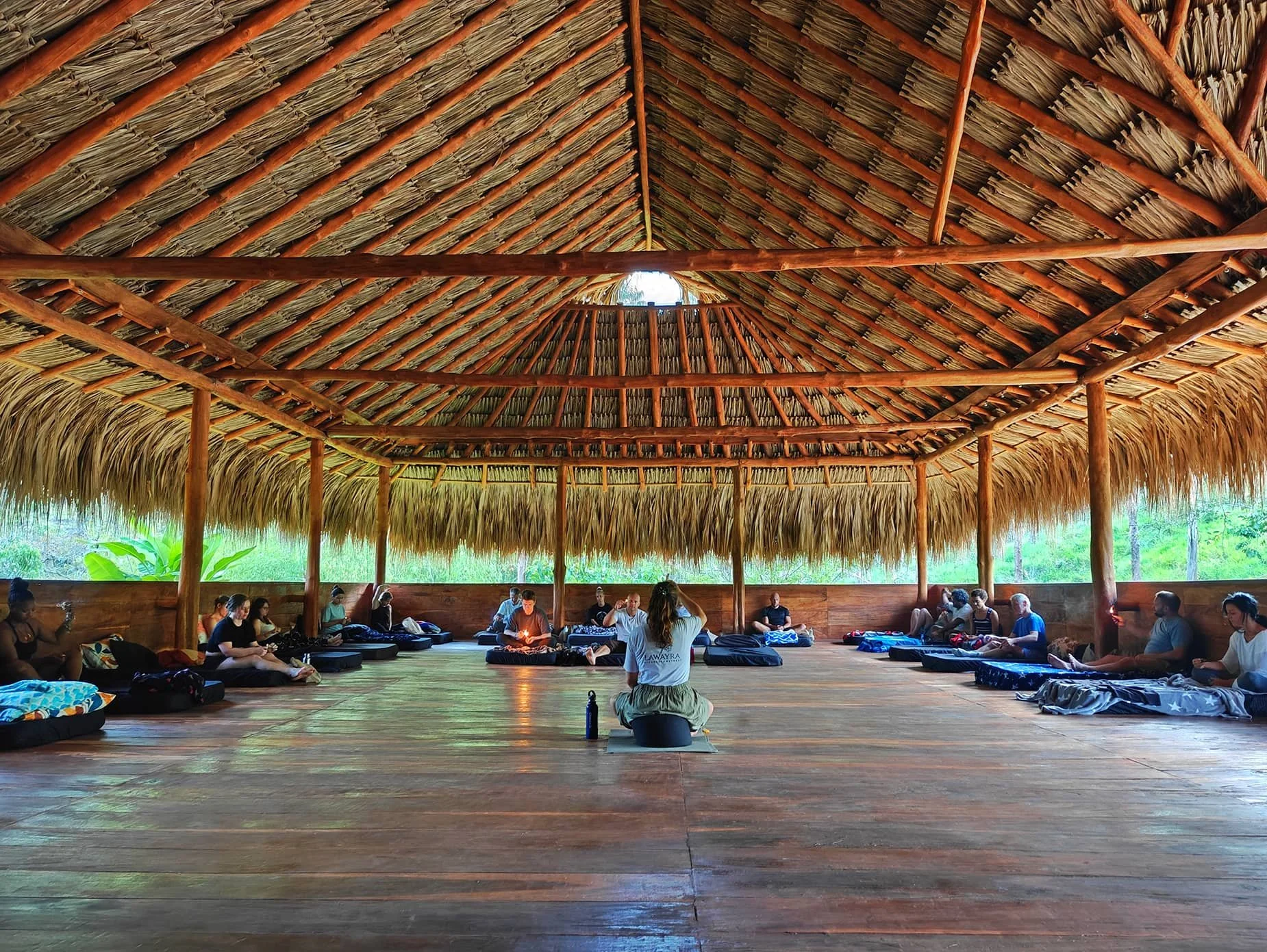 People sitting on mats in a circle inside a thatched roof hut, participating in a group activity or meditation.