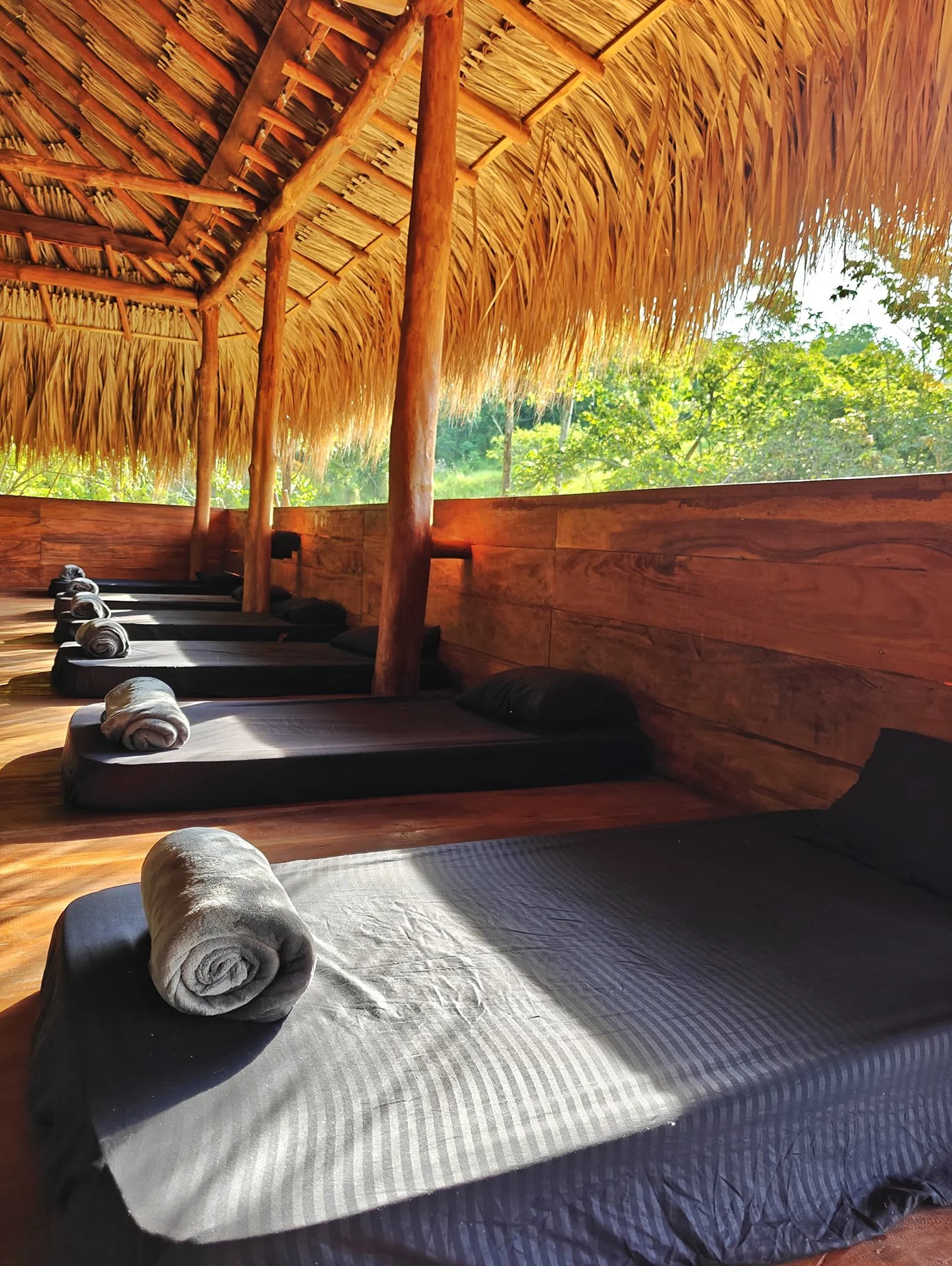 Beachside outdoor massage area with several massage beds covered in gray sheets and rolled white towels, set under a thatched roof with bamboo poles, overlooking lush green trees outside.