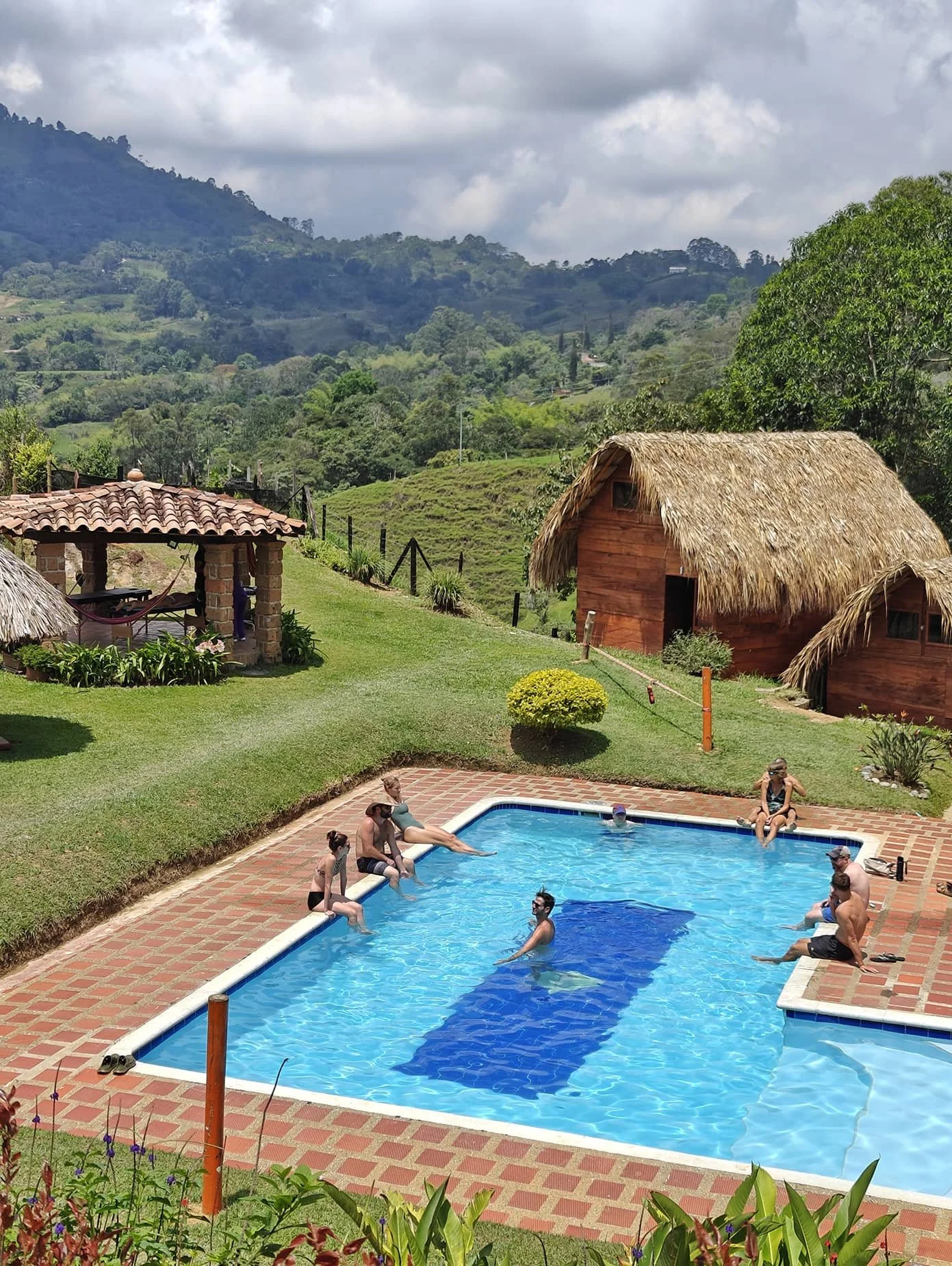Family swimming and relaxing in a backyard pool with scenic green hills and thatched-roof structures in the background.