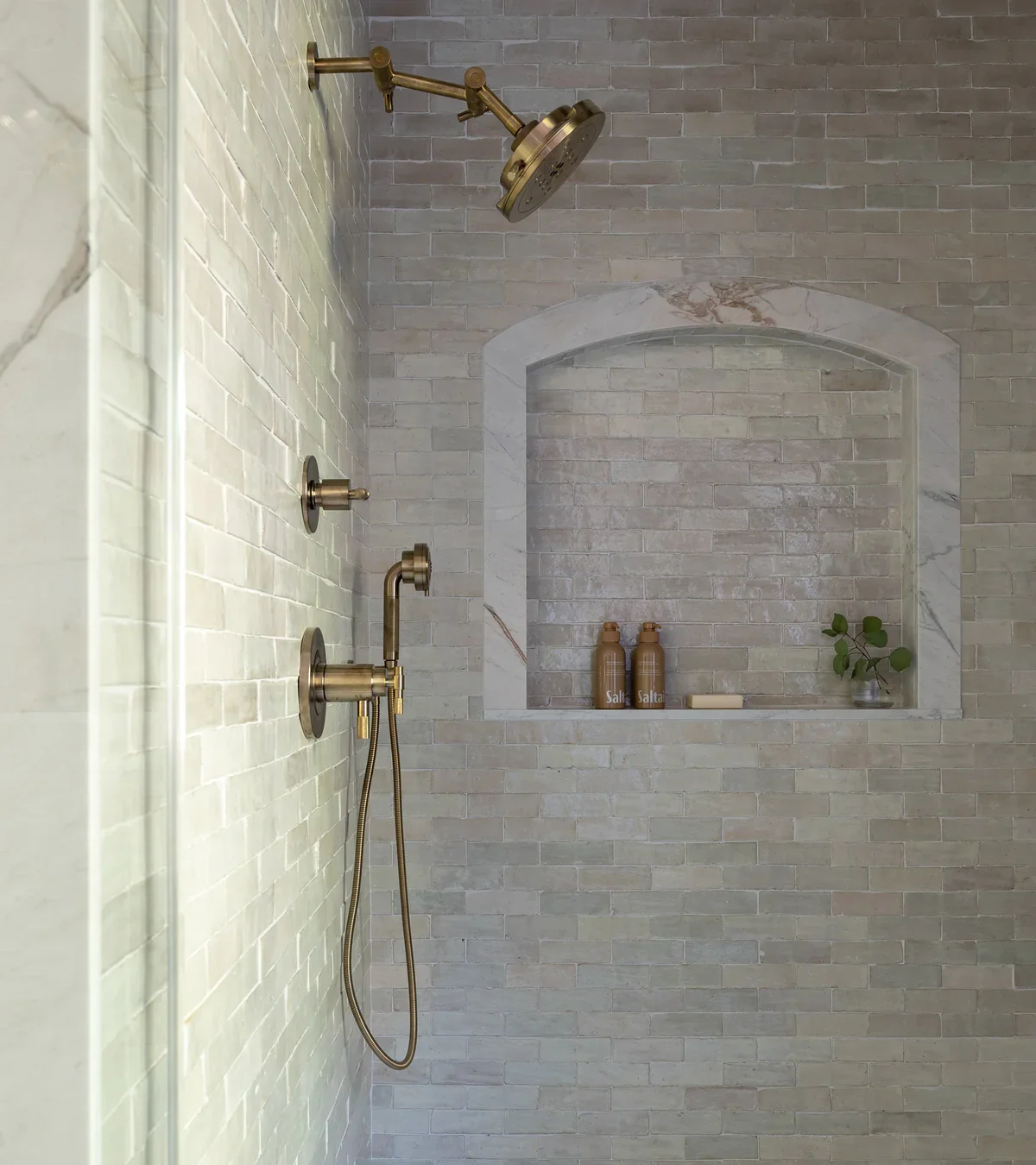 Shower area with beige brick tiles, a gold showerhead, and a built-in niche with bottles of salt and a small plant.