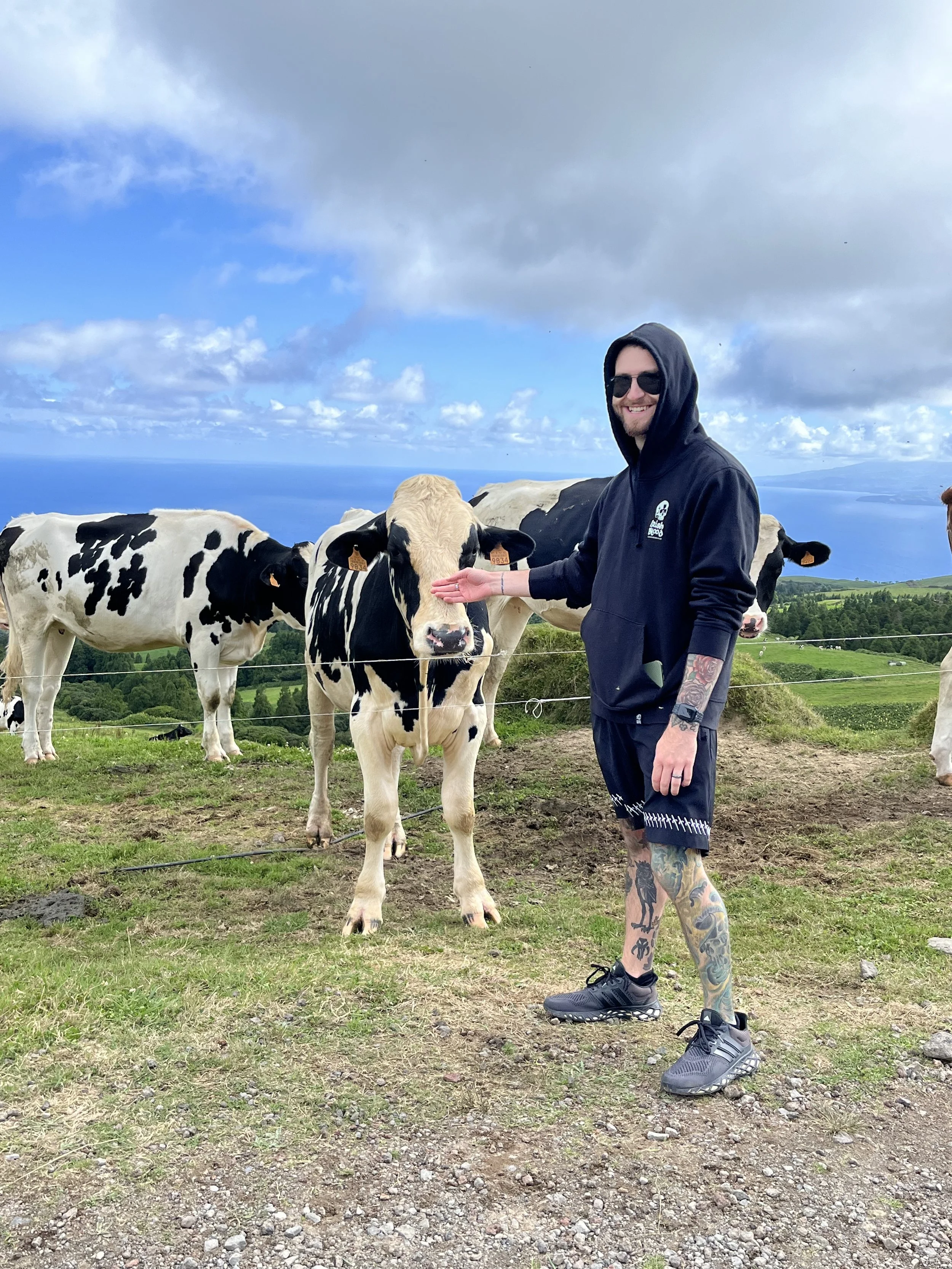 A man wearing sunglasses, a black hoodie, and black shorts with tattoos on his legs is smiling and reaching out to a black and white cow in a farm setting with green hills, a fence, and a cloudy sky in the background.