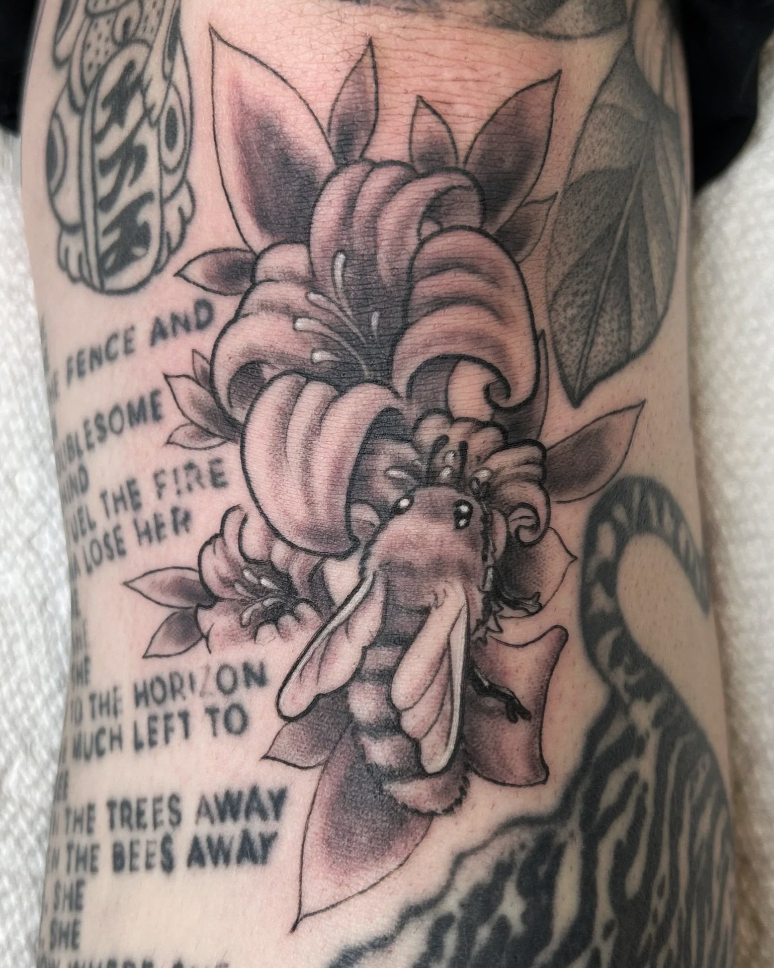 A black and gray tattoo of a large flower with leaves on a person's skin, surrounded by text that is partially visible.