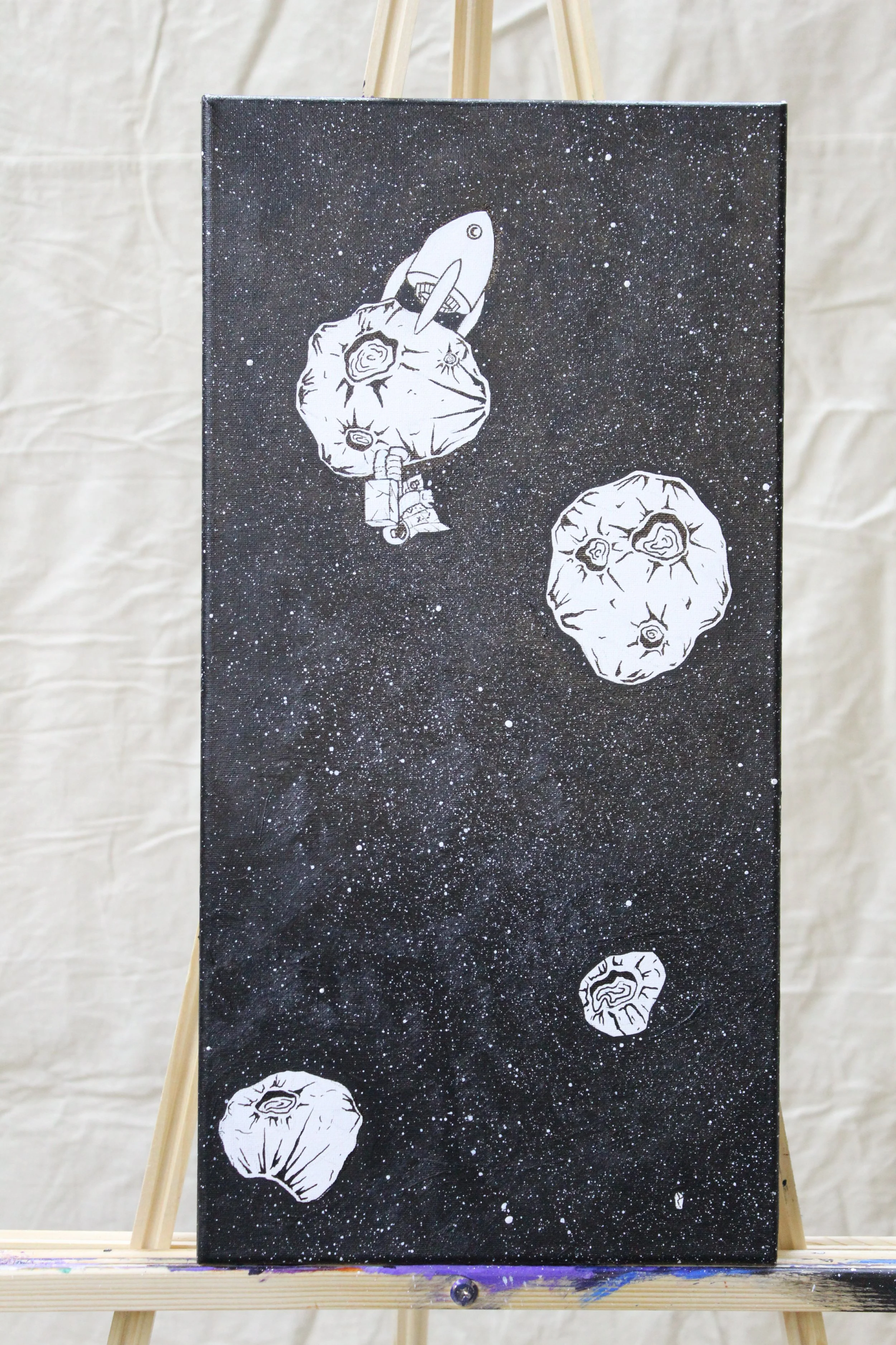 Black canvas with white illustrations of asteroids and a rocket ship in space, displayed on an easel.