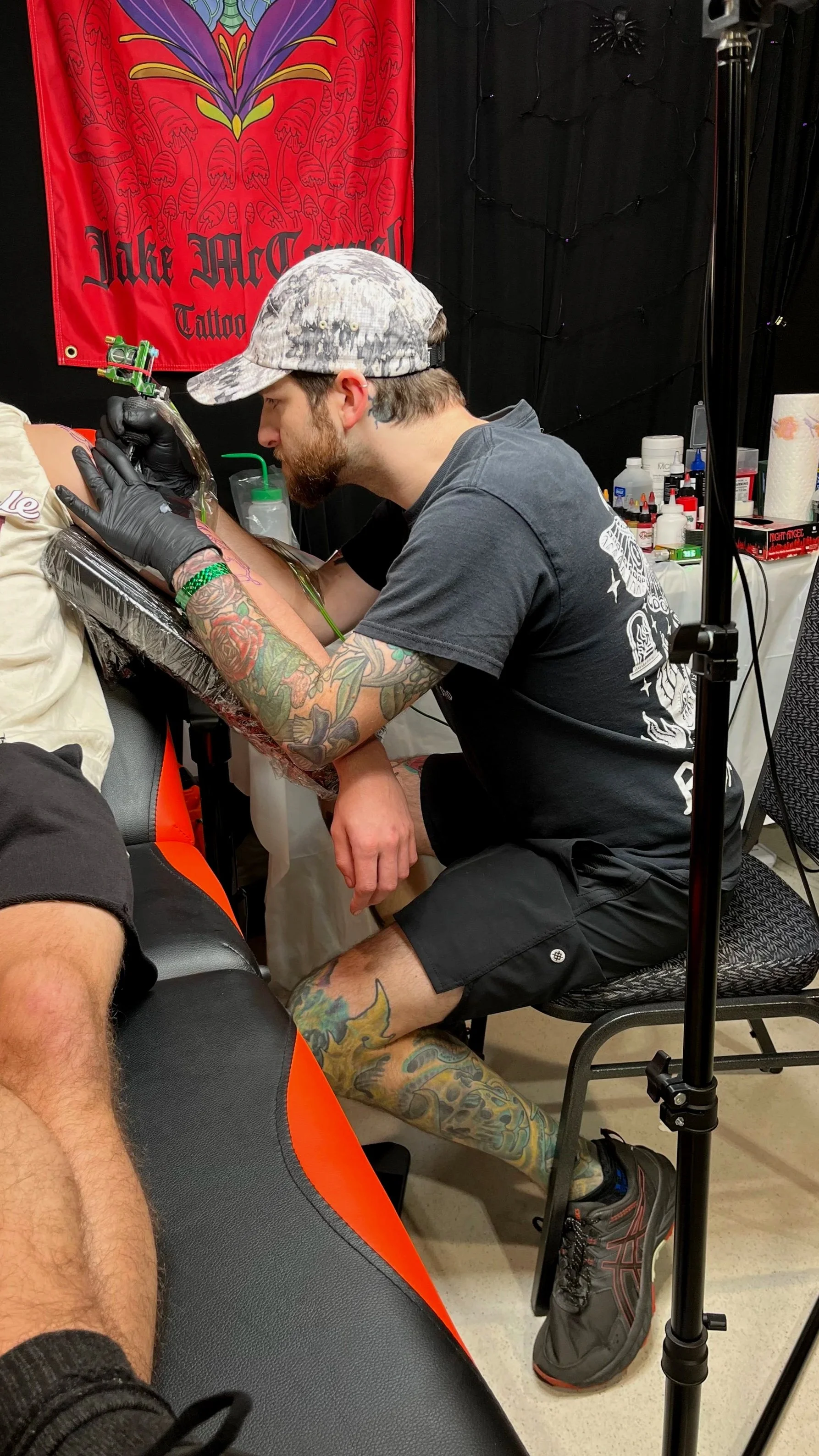 A tattoo artist wearing a baseball cap tattooing a person's arm in a tattoo studio. The studio has a black wall with string lights, various tattoo supplies on a table, and a red banner with a butterfly and flamingo design.