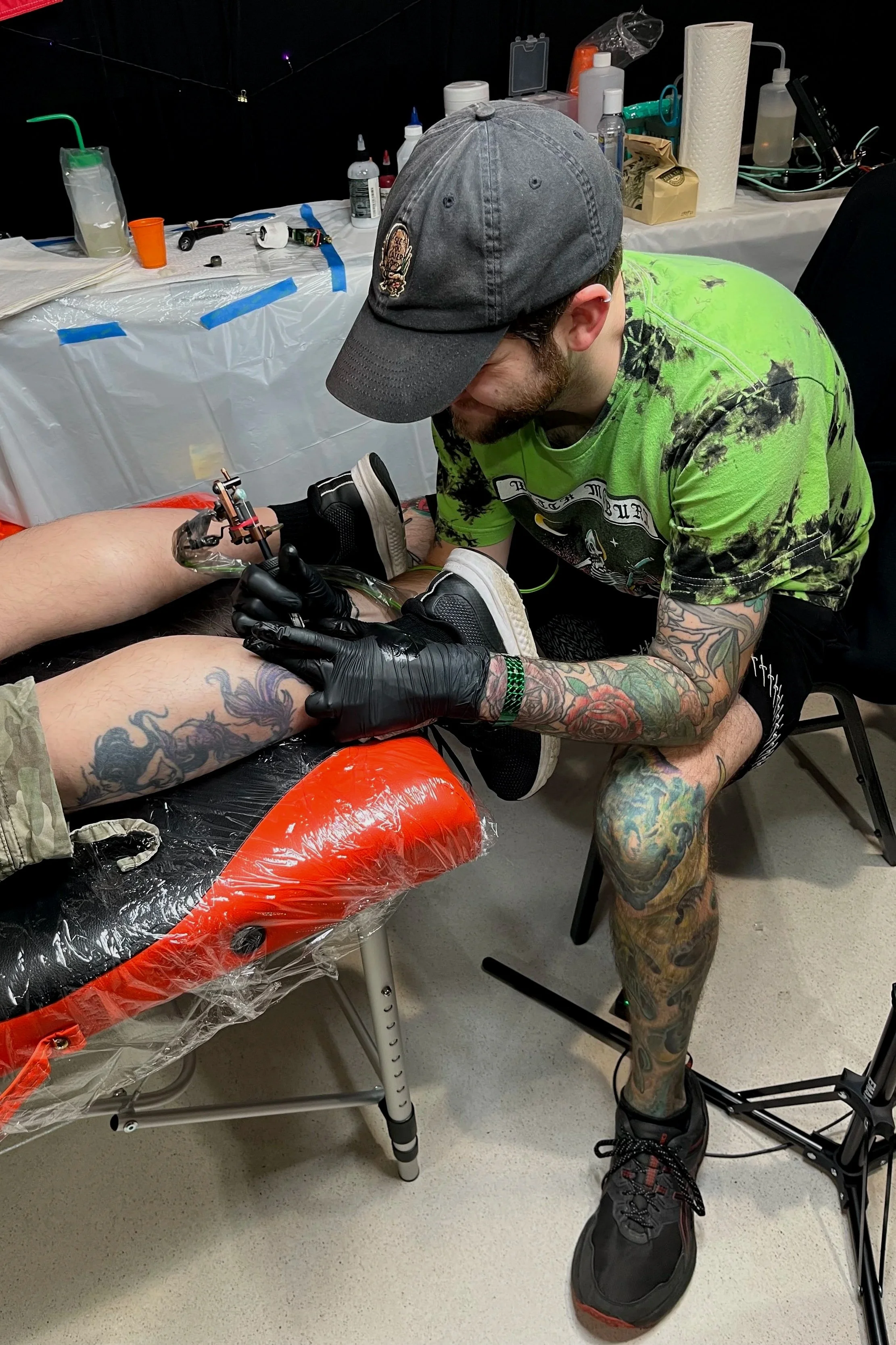 A man with colorful tattoos on his left leg and arm, wearing black sneakers, a green tie-dye t-shirt, a gray cap, and black gloves, tattooing a person's leg in a tattoo studio. The studio has a plastic-covered work table with tattoo supplies in the background.