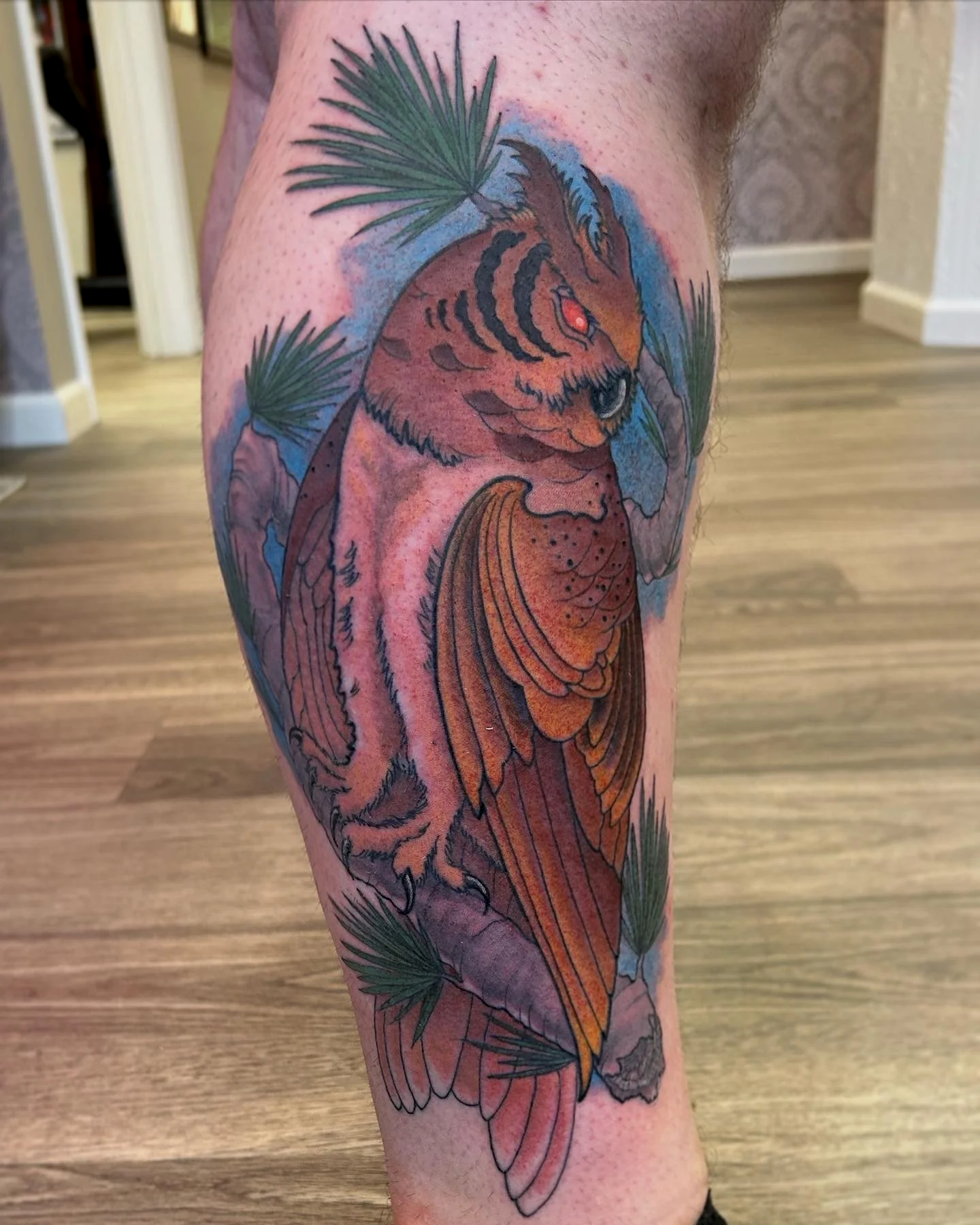 Colorful tattoo of an owl surrounded by green pine needles on a person's arm.