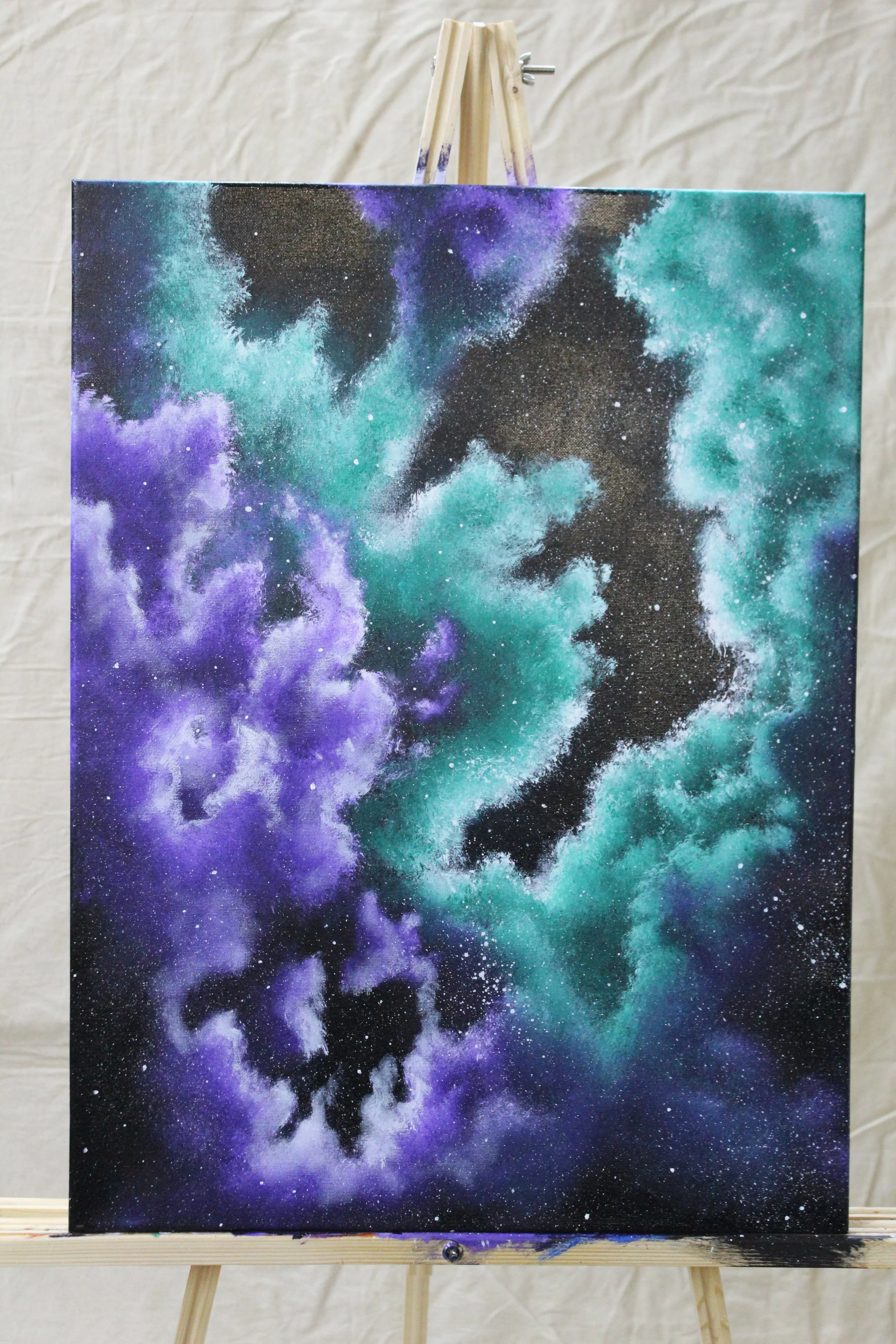 A painting of a galaxy with purple, teal, and black nebula clouds on a canvas displayed on an easel.