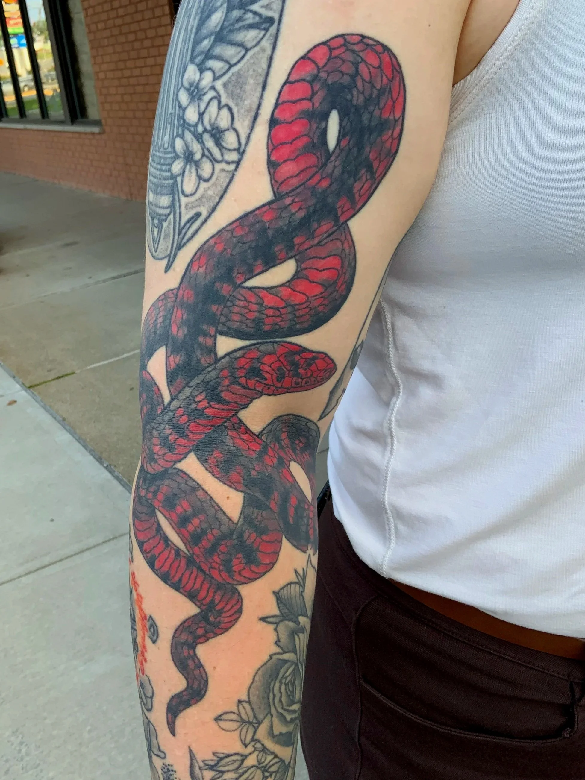 Tattoo of a red and black snake with flowers on the arm of a person wearing a sleeveless white shirt and dark pants.