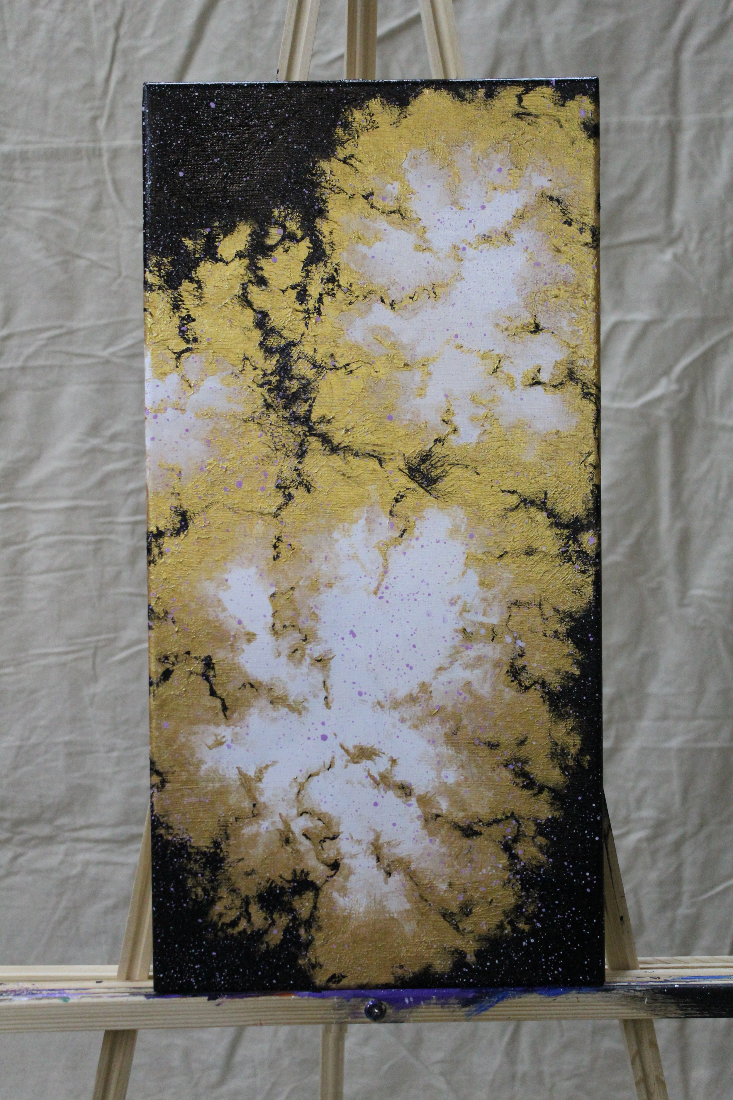 Abstract painting with black, white, and gold splatters on a canvas, displayed on an easel.