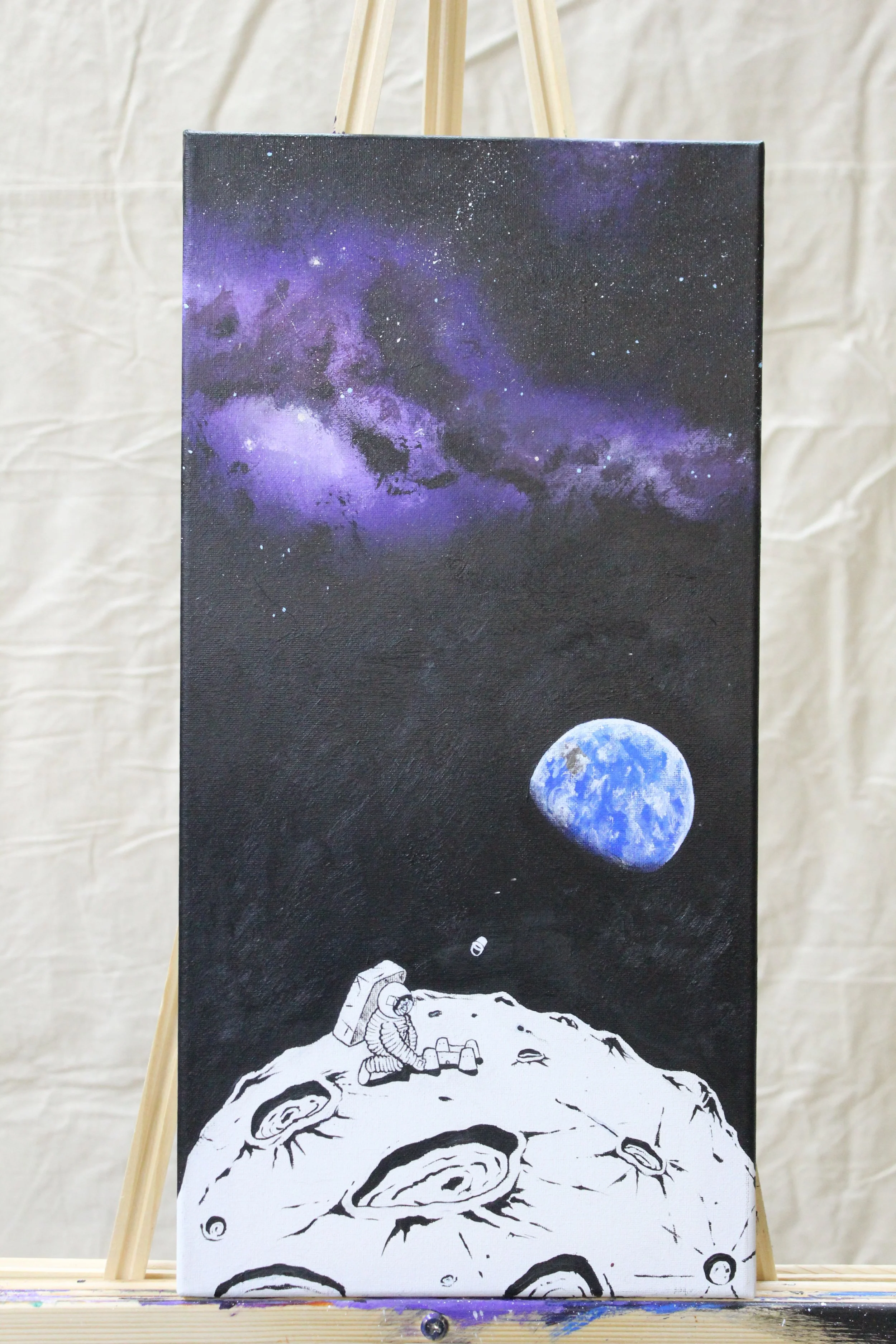 A painting on a canvas depicting an astronaut sitting on the moon with Earth visible in space, featuring a starry galaxy with purple and black hues in the background.