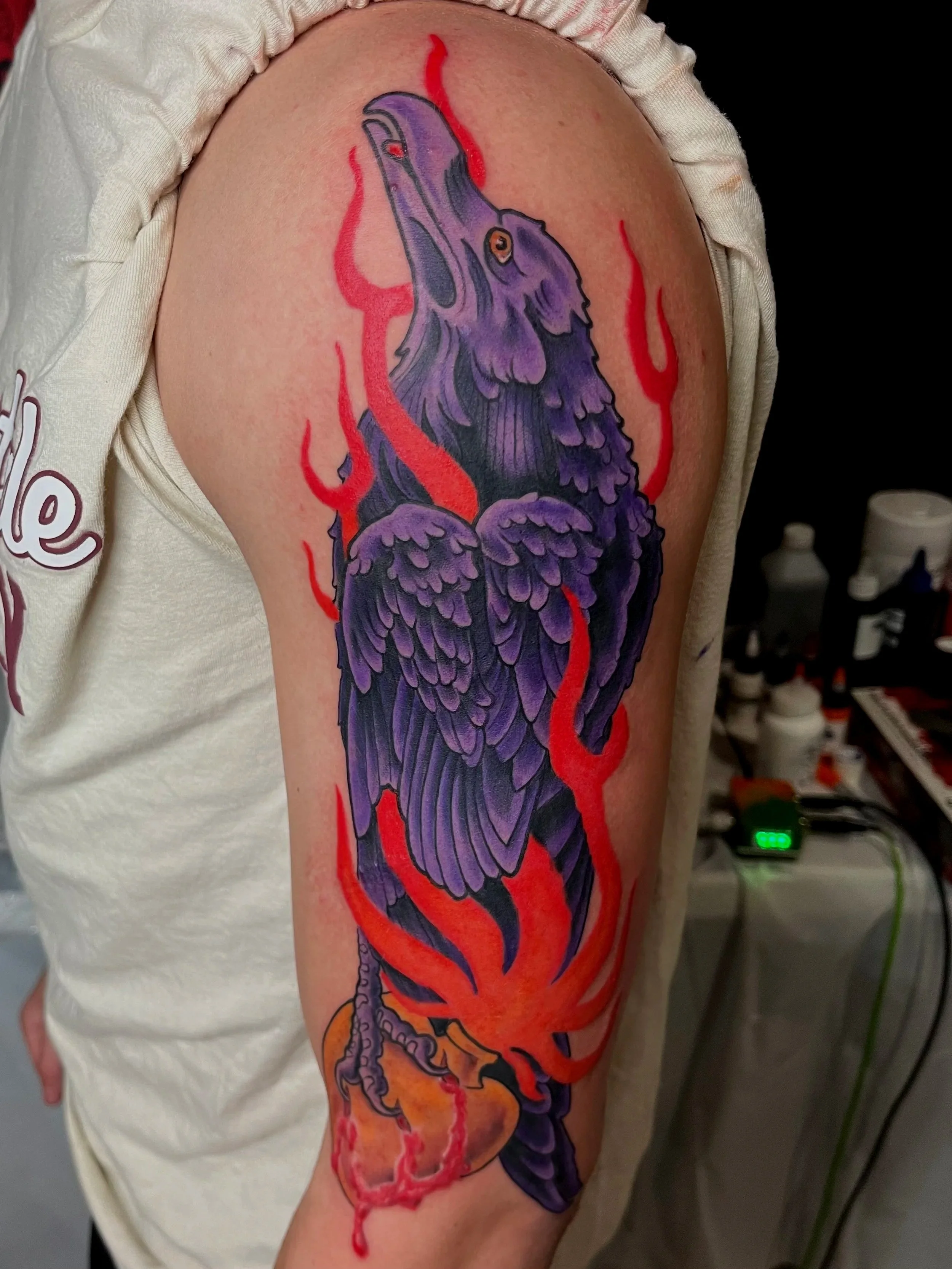 A colorful tattoo of a purple raven with red flames on the shoulder.