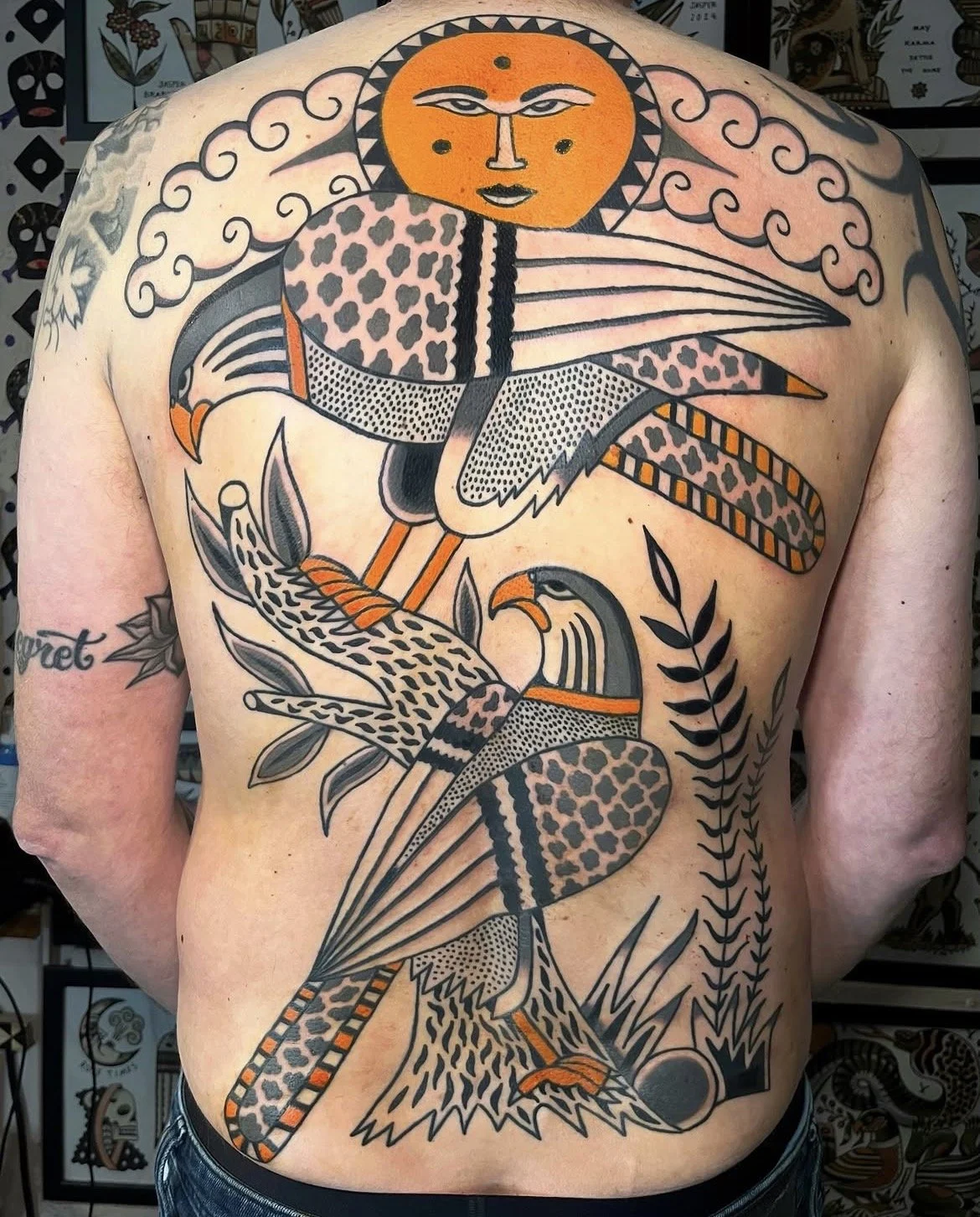A traditional-style tattoo on a person's back, featuring a large bird perched on a branch with a leopard pattern body, surrounded by cloud and plant motifs, and a sun with a face at the top.