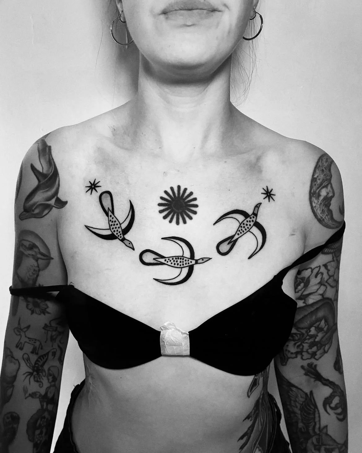 A woman with tattoos on her arms and chest, wearing a black bra and hoop earrings, standing against a plain wall.