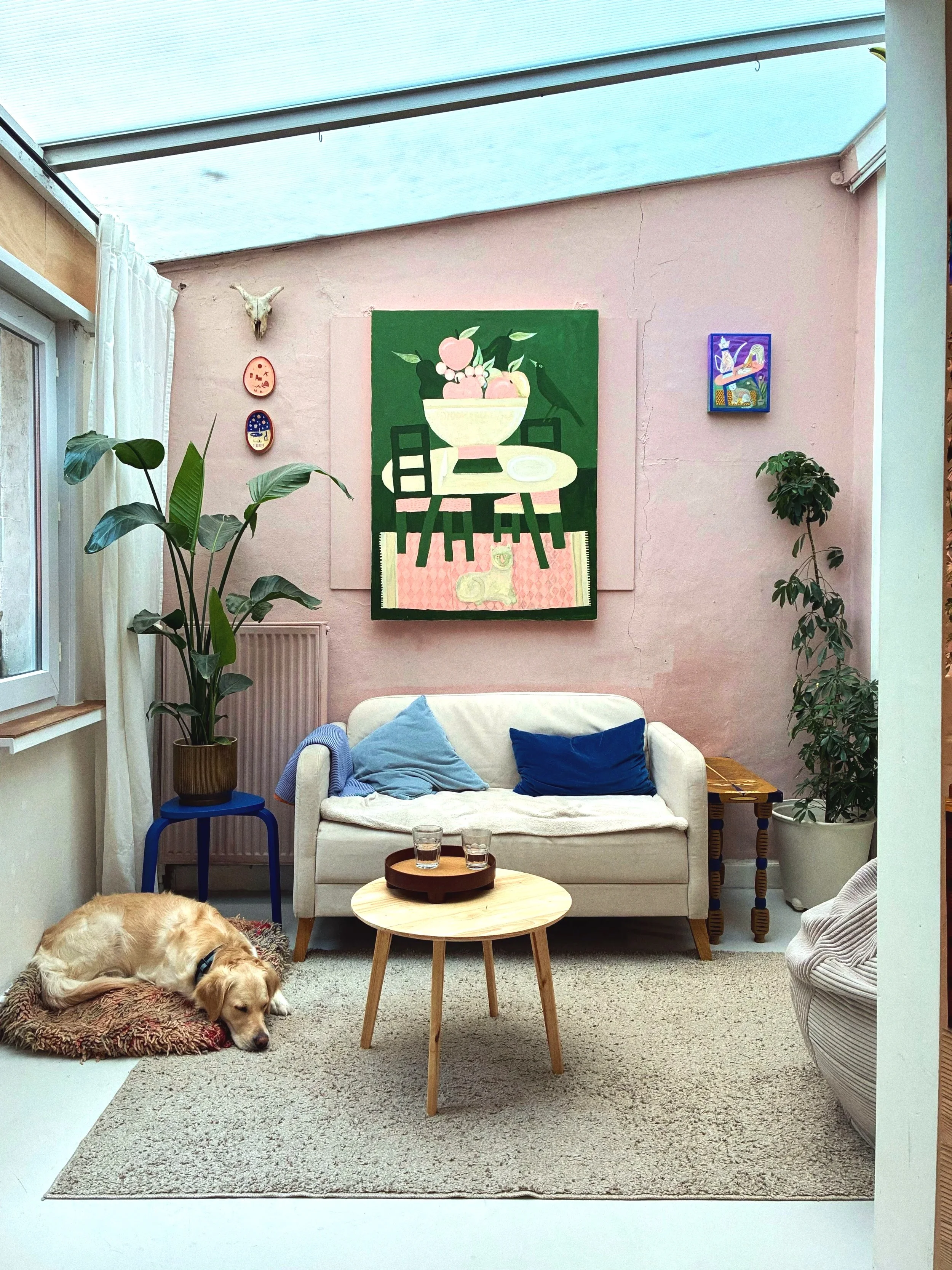 A cozy living room with light pink walls and a skylight ceiling. There is a white loveseat with blue pillows, a small round wooden coffee table with two glasses, and a Golden Retriever dog sleeping on a rug. Decor includes artwork on the walls, a pot