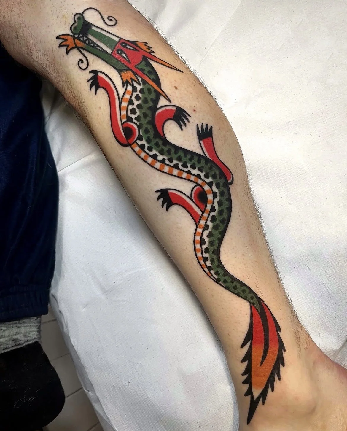 Colorful tattoo of a stylized dragon with green, red, orange, and black details on a person's calf.