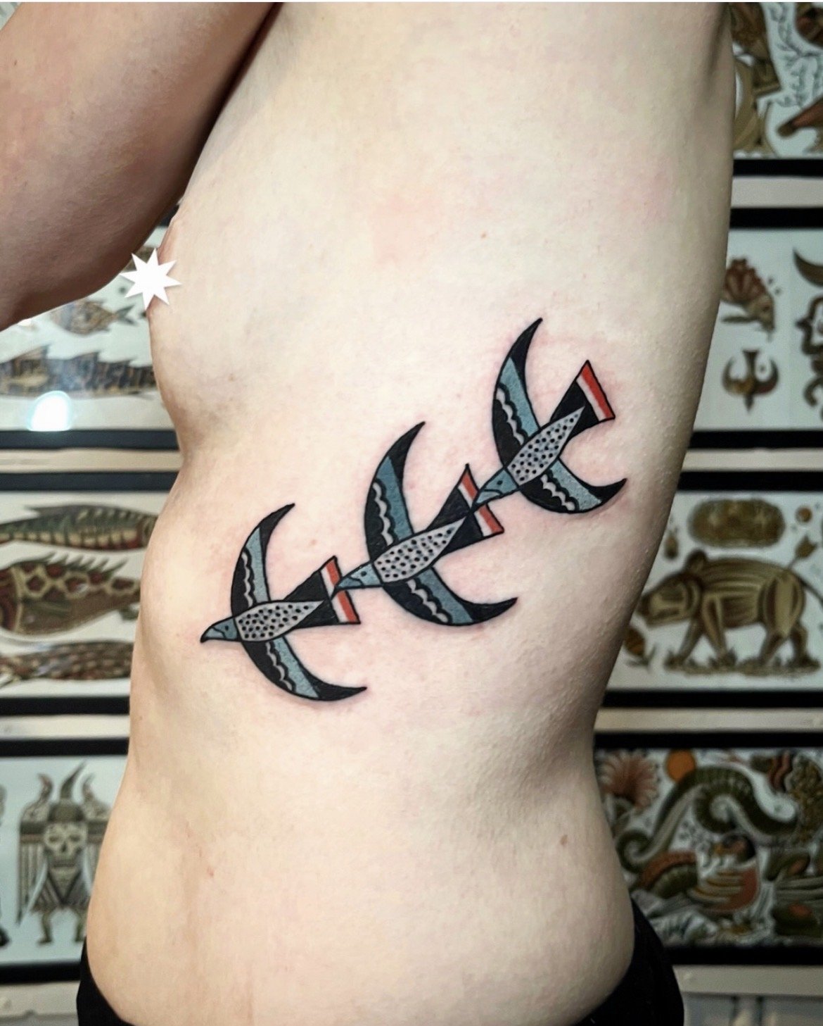 Tattoo of a stylized fish skeleton with black, gray, red, and blue coloring on a person's side.