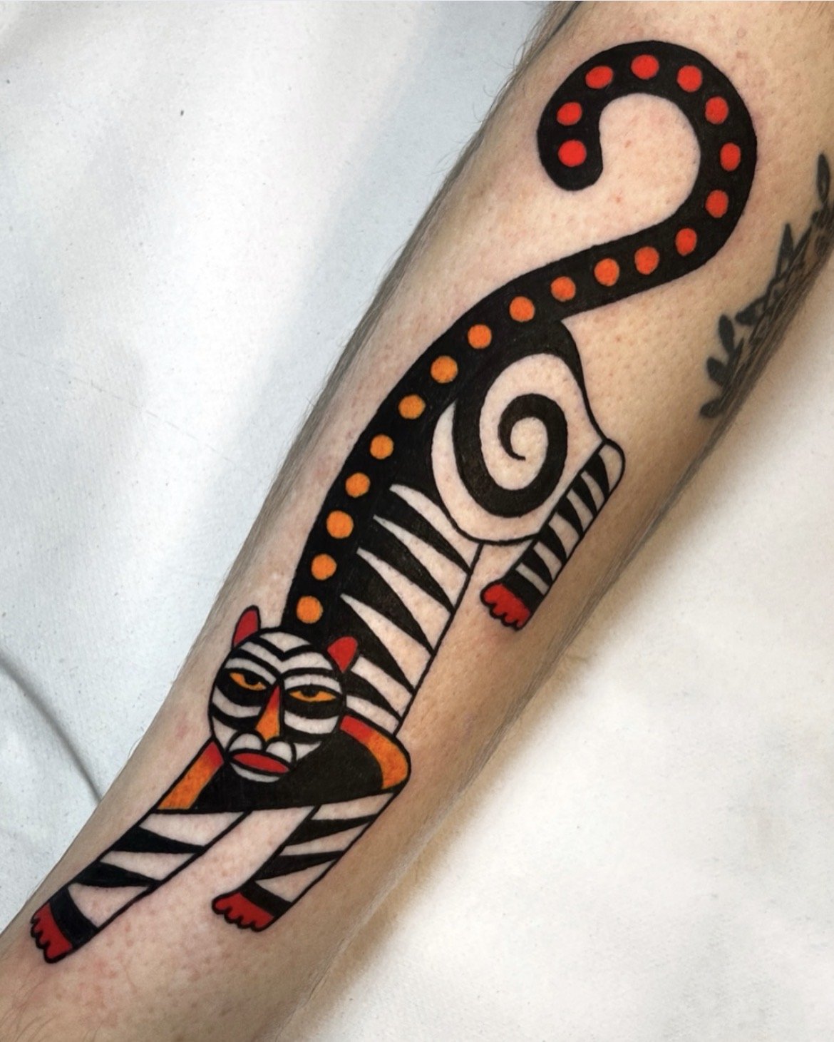Tattoo of a stylized cat with black and white stripes and red accents, featuring a spiral tail and a face with yellow eyes.