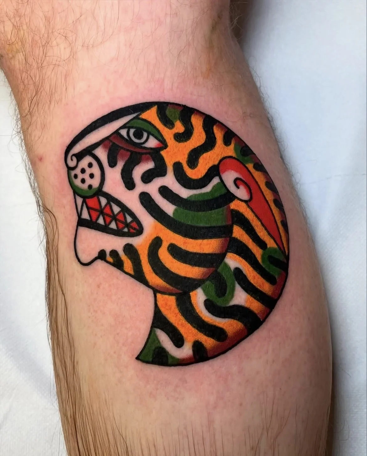 Colorful tattoo of a stylized face with an eye, open mouth, and swirling patterns in orange, green, red, and black, on a person's arm.