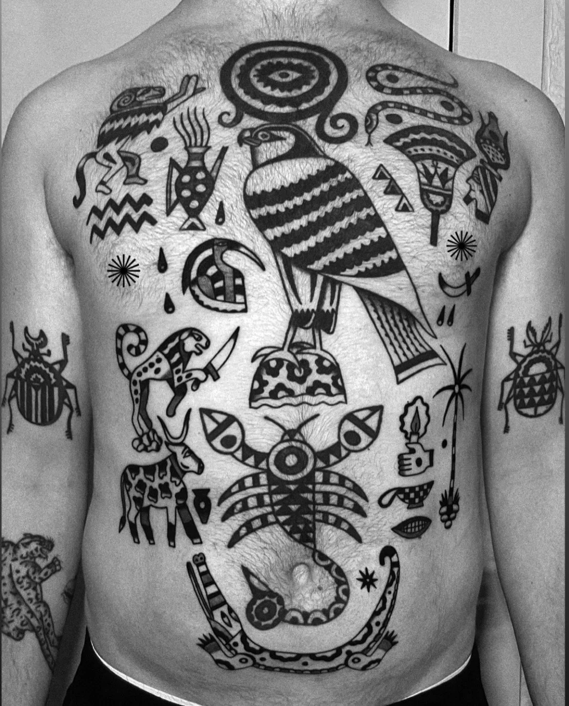 Black and white photograph of a person's chest and abdomen covered with various traditional-style tattoos, including a large eagle, snakes, insects, and decorative patterns.