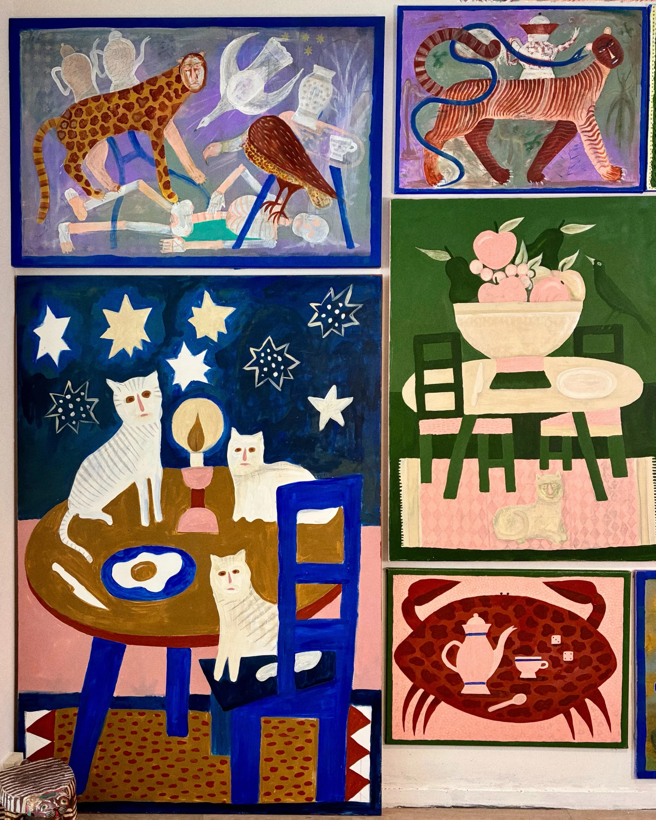 A colorful art display featuring multiple paintings with animal and abstract themes. The paintings include a leopard with a human face attacking a human figure, a tiger with a human face and a snake on its back, a white tiger with a golden halo surrounded by stars, and a green background with a table holding a bowl of fruit, a teapot, and cups.