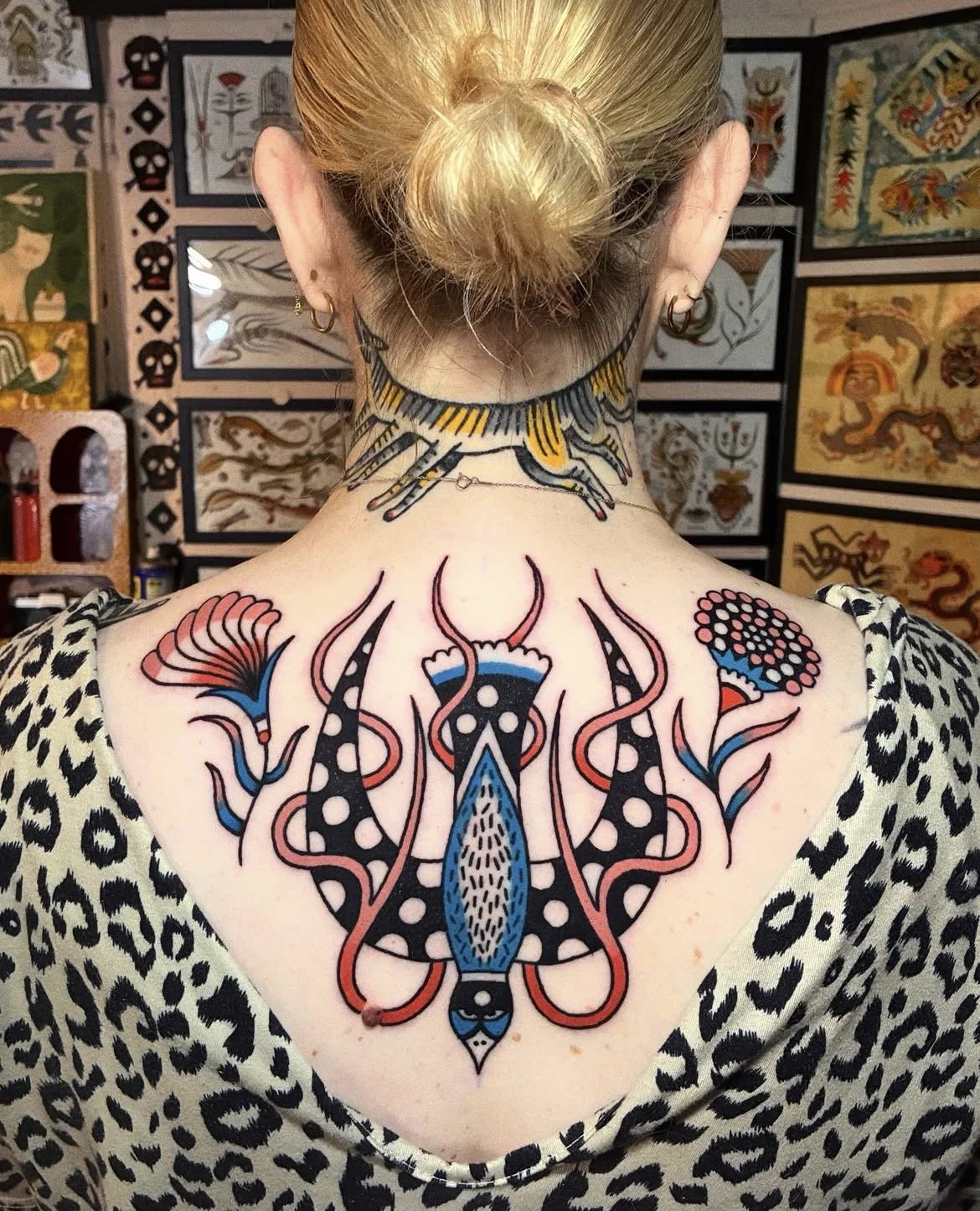 Colorful tattoos on a woman's back and neck featuring abstract and floral designs in black, red, blue, and white, with a background of tattoo artwork on display.