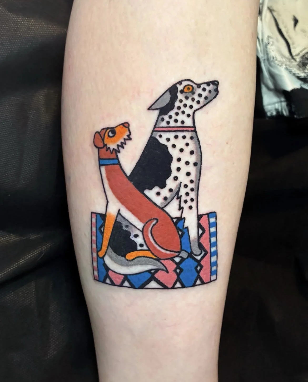 Tattoo of two dogs, one dog with a Dalmatian pattern sitting upright, and another dog with orange and black coloring sitting on a patterned mat.