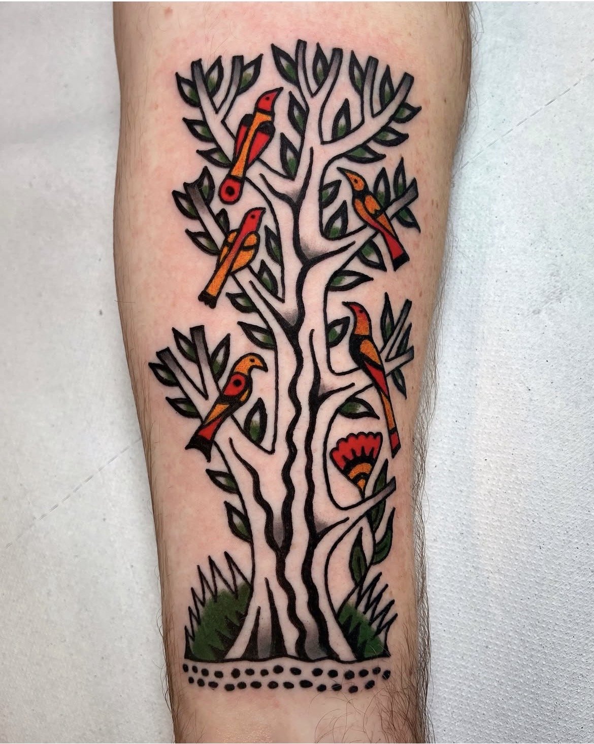 Tattoo of a stylized tree with black outlines, green leaves, red, yellow, and orange birds, and a red and black flower at the base.