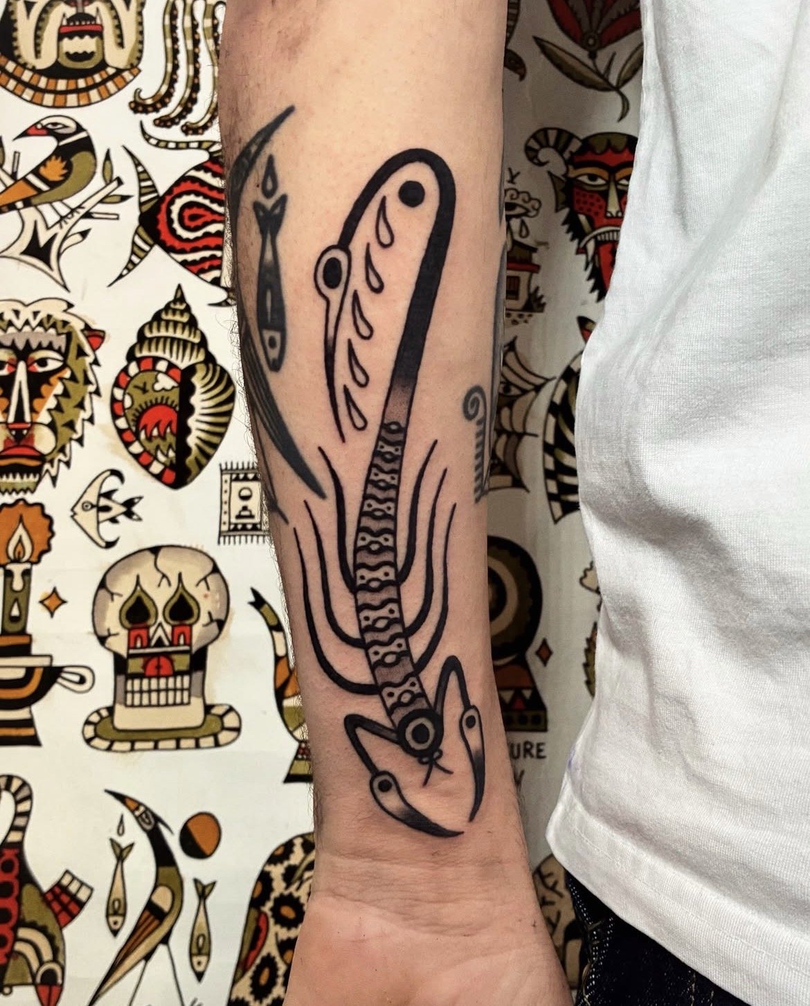 Black line tattoo of a fish or aquatic creature on a person's forearm, with a patterned tail and stylized features.