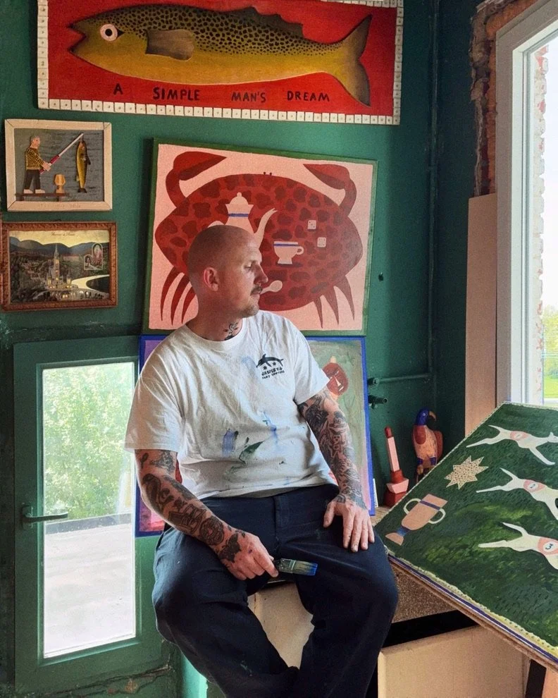 A young man with tattoos sitting on a stool inside an art studio, surrounded by colorful paintings of animals, including a fish, a crab, and a herd of cows. Sunlight streams in from a window on the right.