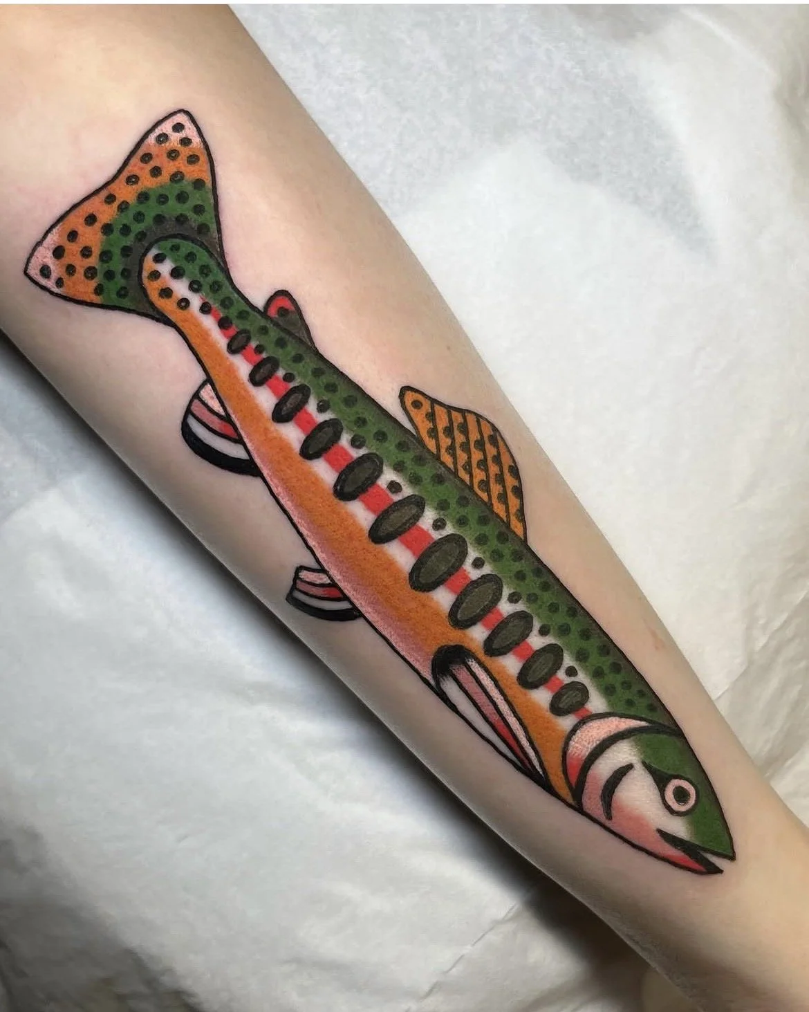 Colorful tattoo of a fish on a person's arm, with green, orange, and black details and dotted patterns.