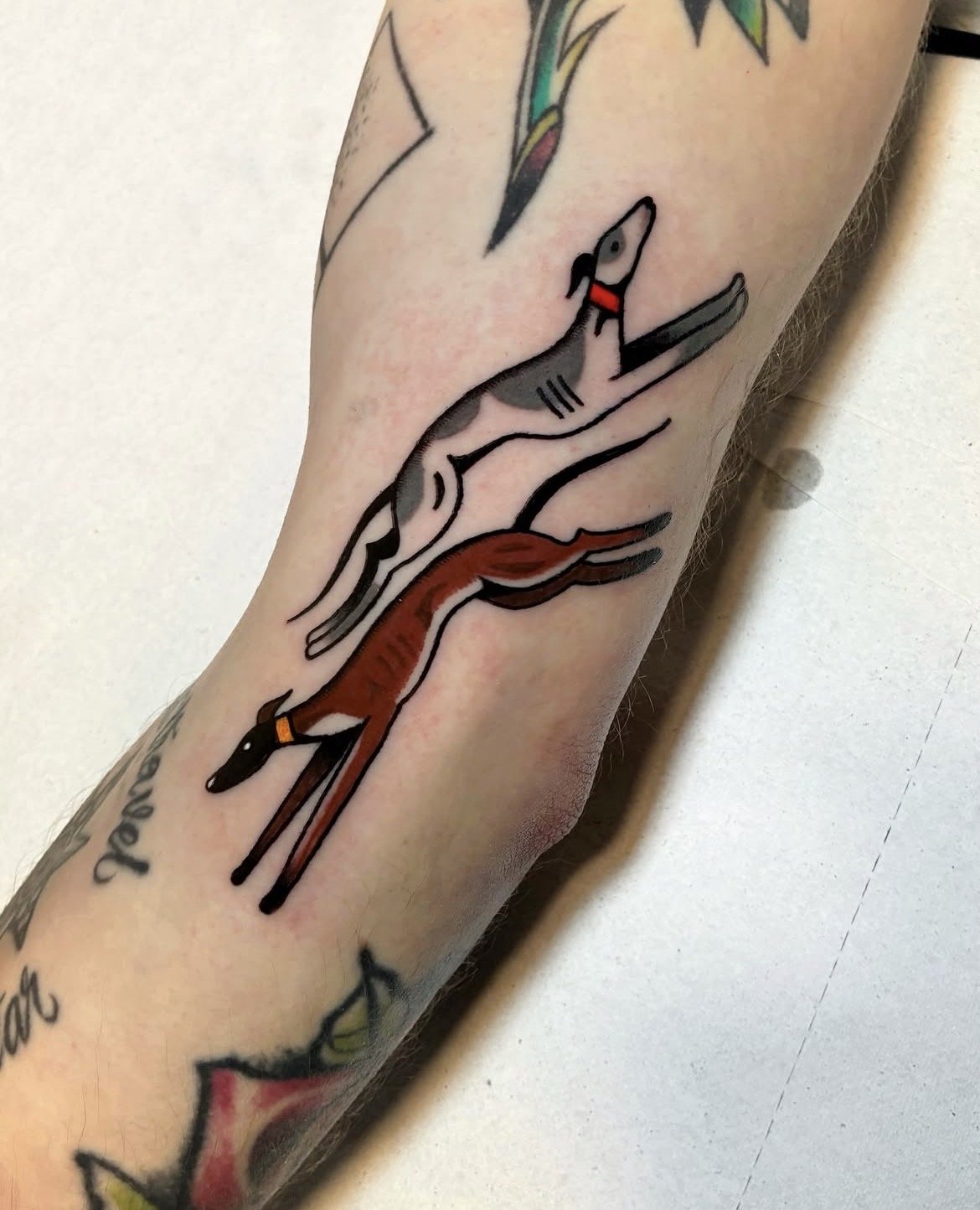 Tattoo of a stylized deer with elongated legs, a red collar, and simple black outlines, located on a person's arm.