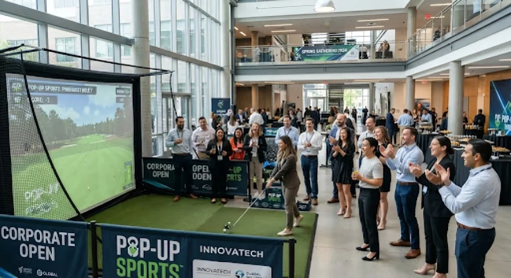 A woman playing golf at an indoor pop-up sports event, with people watching and clapping around her in a modern, glass-walled building.