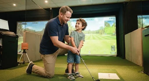 A man and a young boy at an indoor golf simulator, with the man kneeling and helping the boy hold a golf club, both smiling.