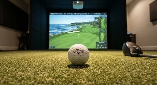 Indoor golf simulation with a golf ball on artificial turf and a large screen displaying a golf course.