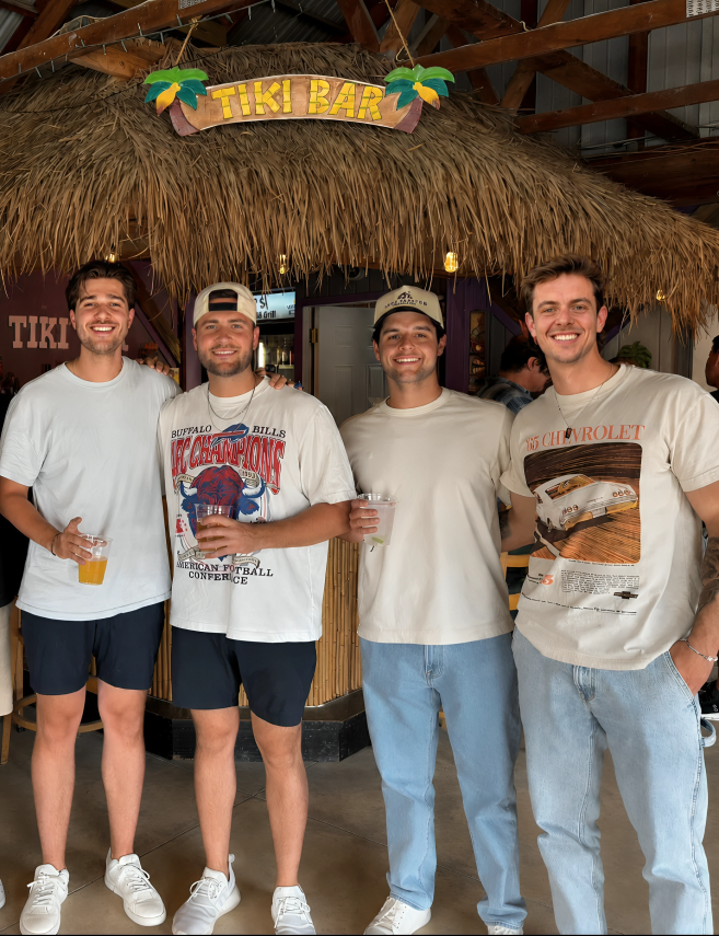 Four young men standing together in front of a tiki bar with a thatched roof, smiling and holding drinks, at a social gathering or party.