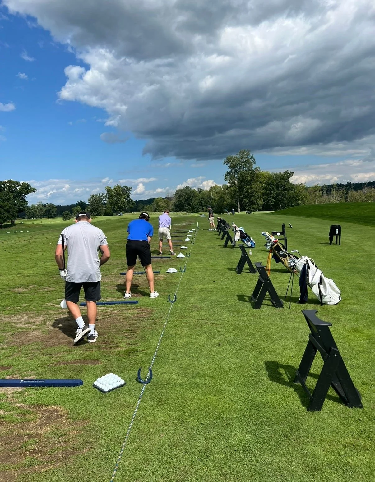 Golfers practicing at a driving range on a grassy field with clouds overhead.