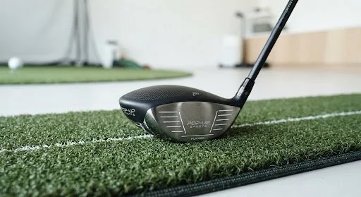 Close-up of a golf driver club on a practice mat with a green turf surface.