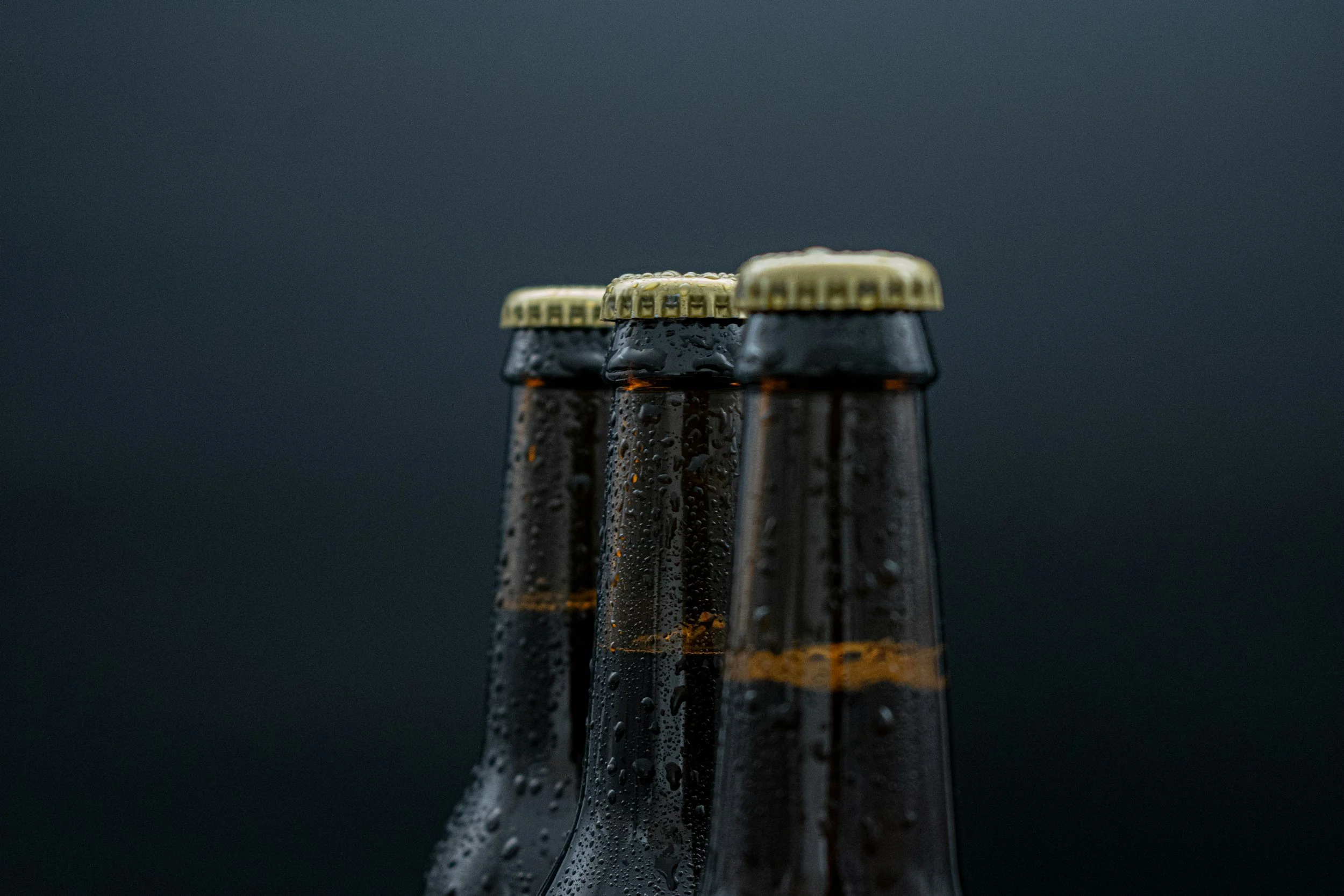 Close-up of beer bottles covered in condensation with caps in focus, against a dark background.