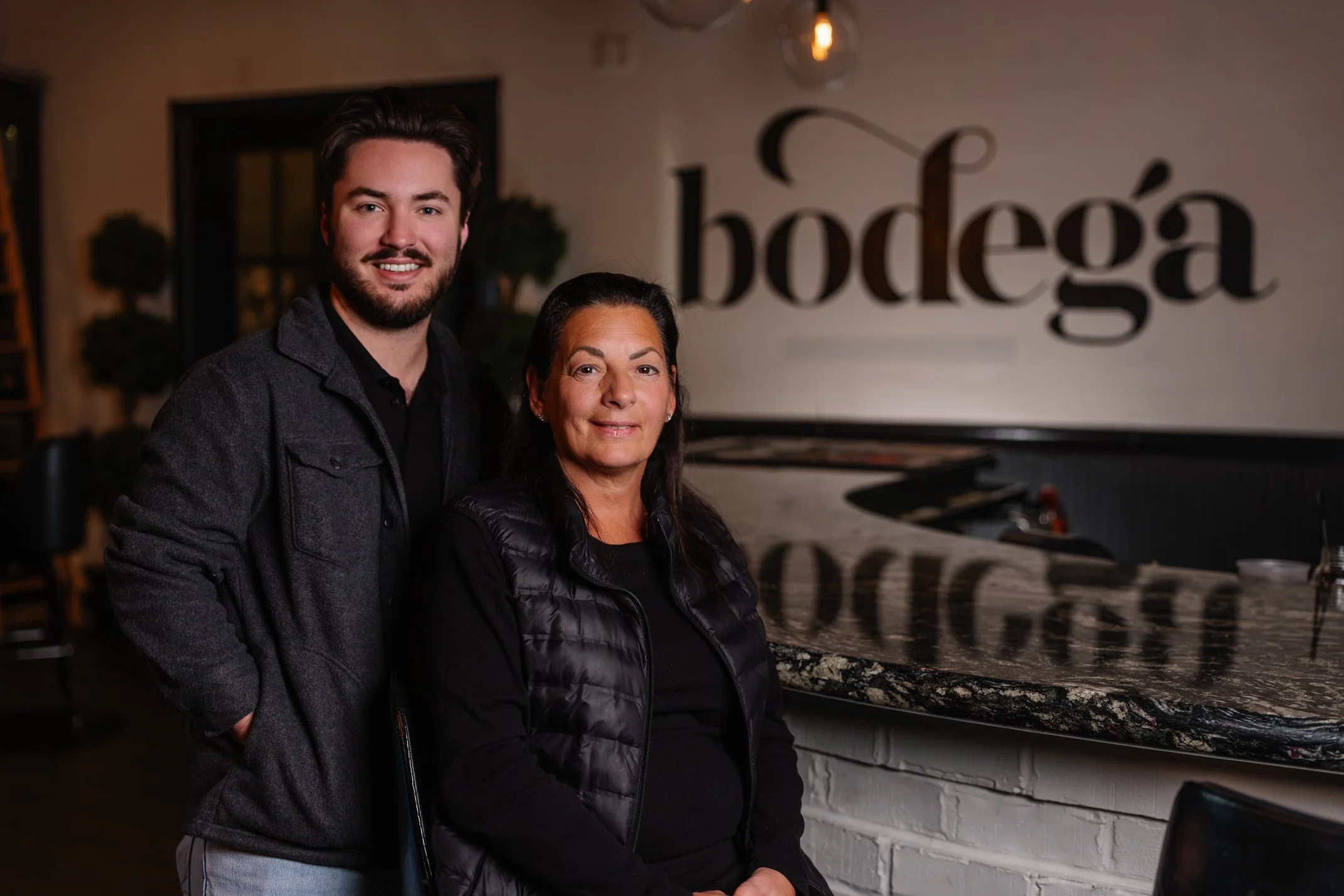 A young man with dark hair and beard, wearing a dark gray jacket, stands behind an older woman with dark hair, wearing a black vest, in a room with a white brick wall and a grand piano. The background has a large sign with the word 'bodega' on the wall.