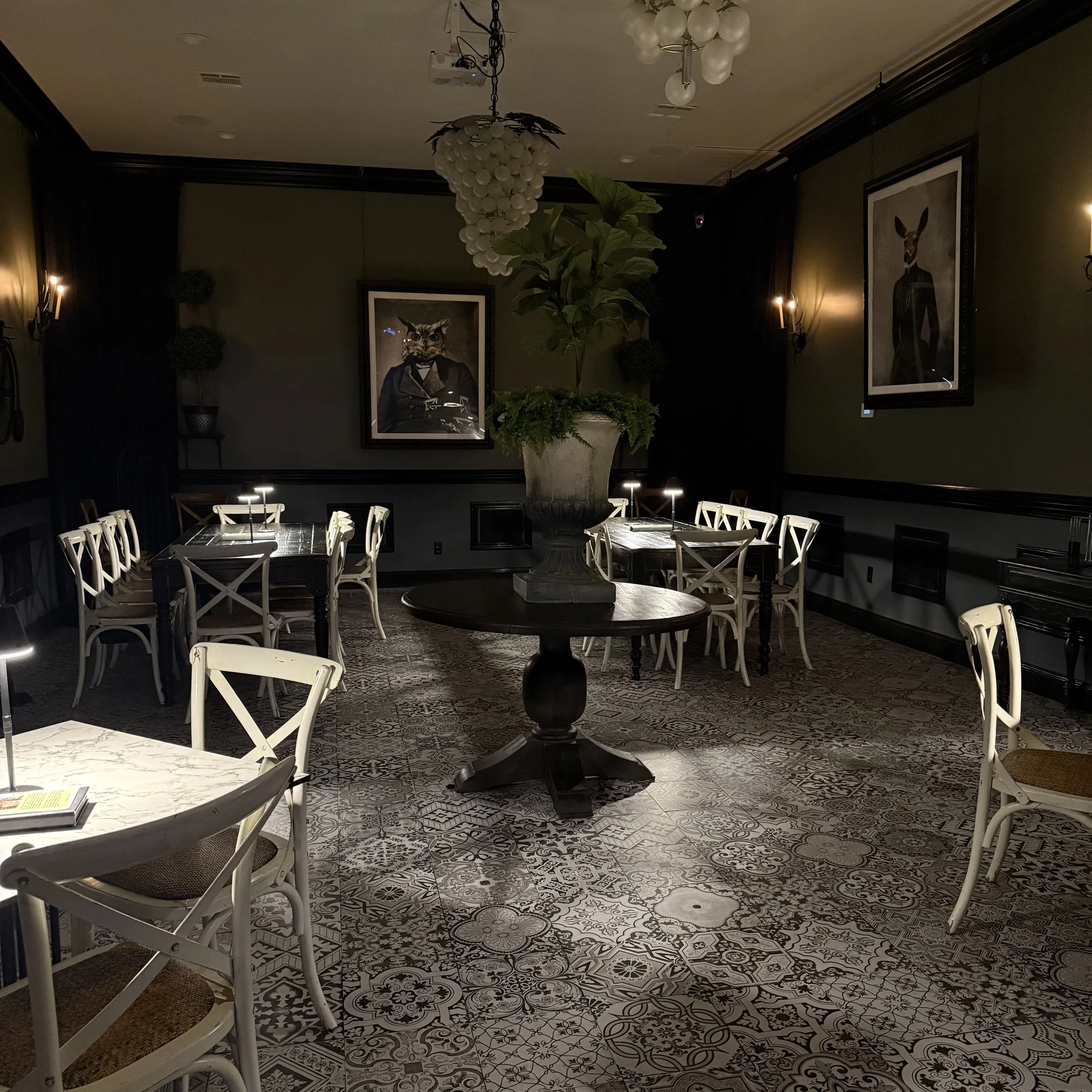 A dimly lit dining room with patterned tile flooring, a large potted plant on a round table, framed artwork on the dark walls, and two chandeliers hanging from the ceiling. The room has white chairs surrounding tables and subtle wall-mounted lighting.