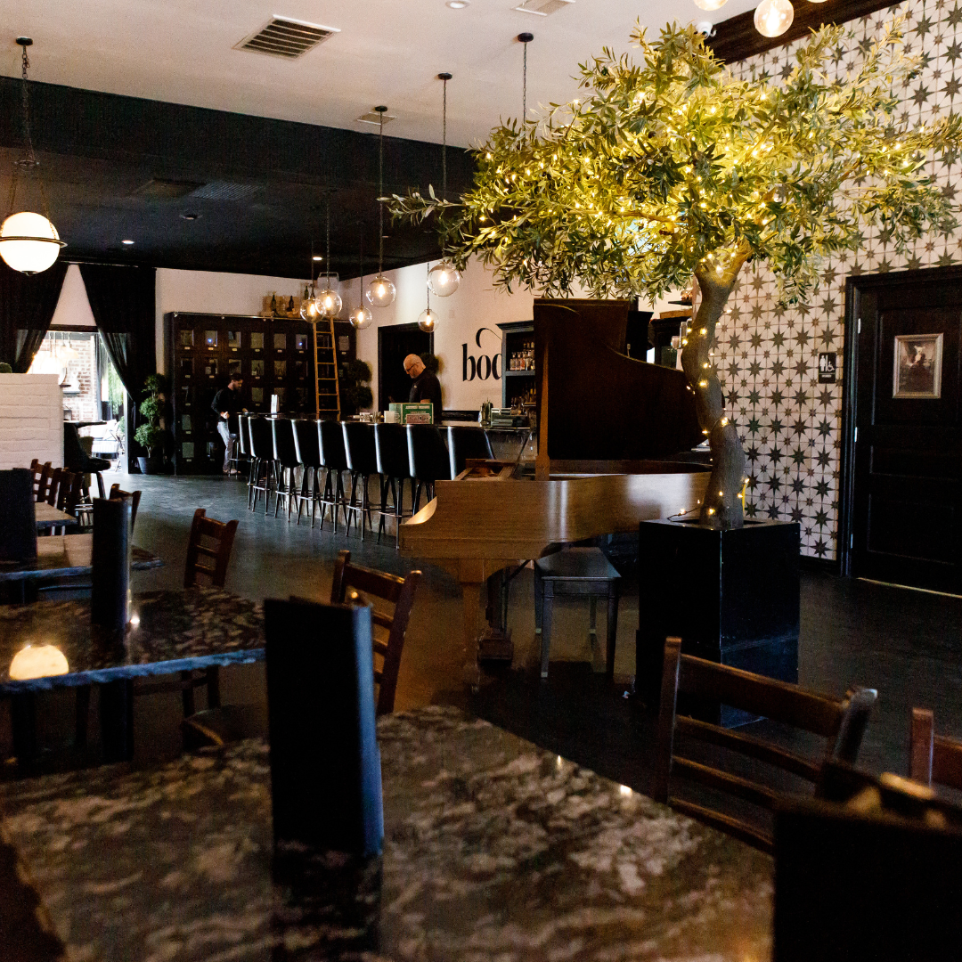 Interior of a cozy restaurant with a bar, wooden furniture, a potted olive tree with fairy lights, and decorative wall patterns.