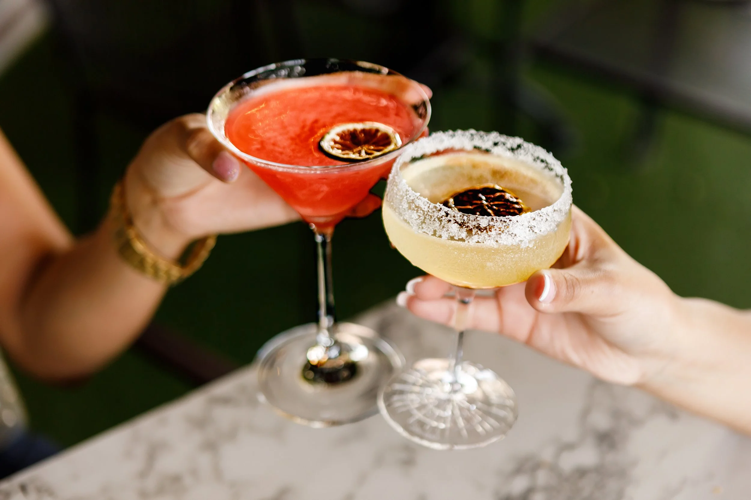 Two hands holding elegant cocktail glasses with salted rims, one glass contains a red-colored drink with a slice of dried citrus, the other has a pale yellow drink with a similar citrus garnish, set against a blurred background.