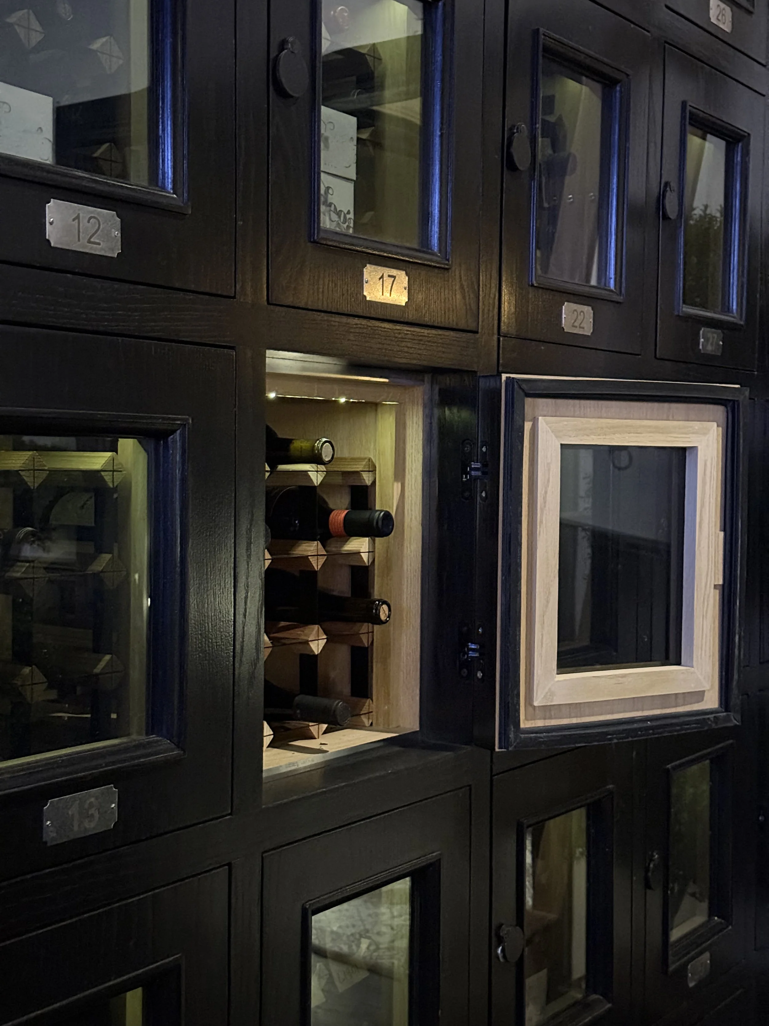 Display of black wooden cabinets with glass doors and numbered silver plaques, containing wine bottles and boxes inside.